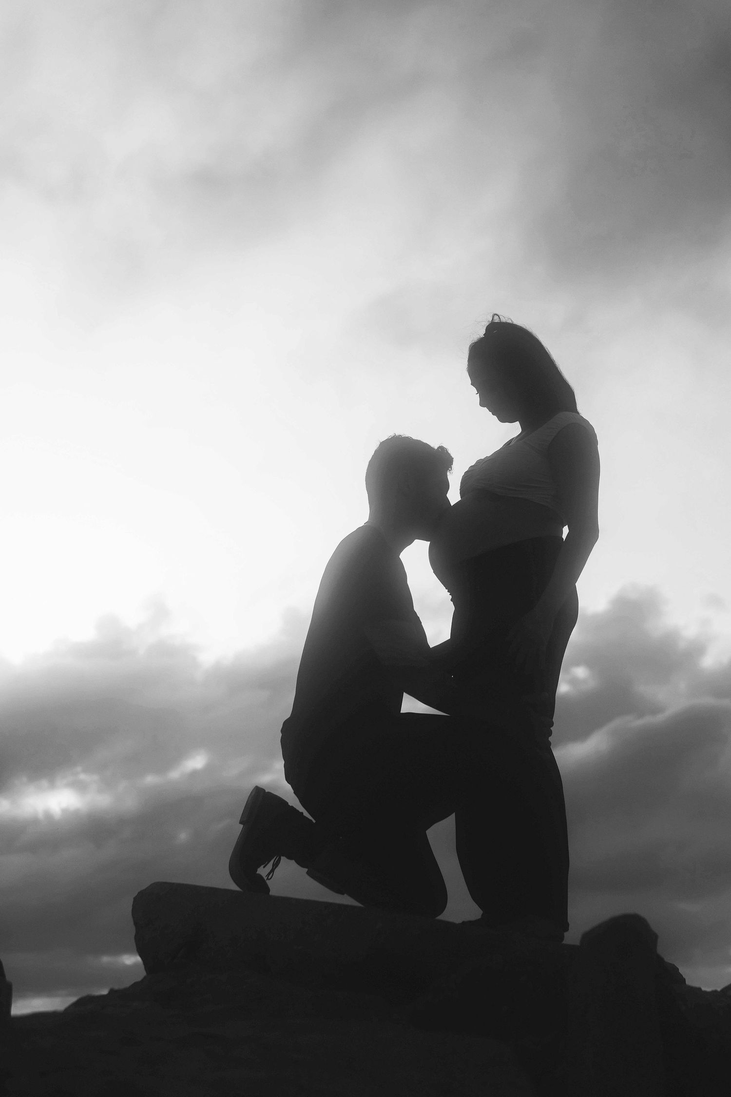 Black and white silhouette of a man kneeling before a pregnant woman against a dramatic cloudy sky.