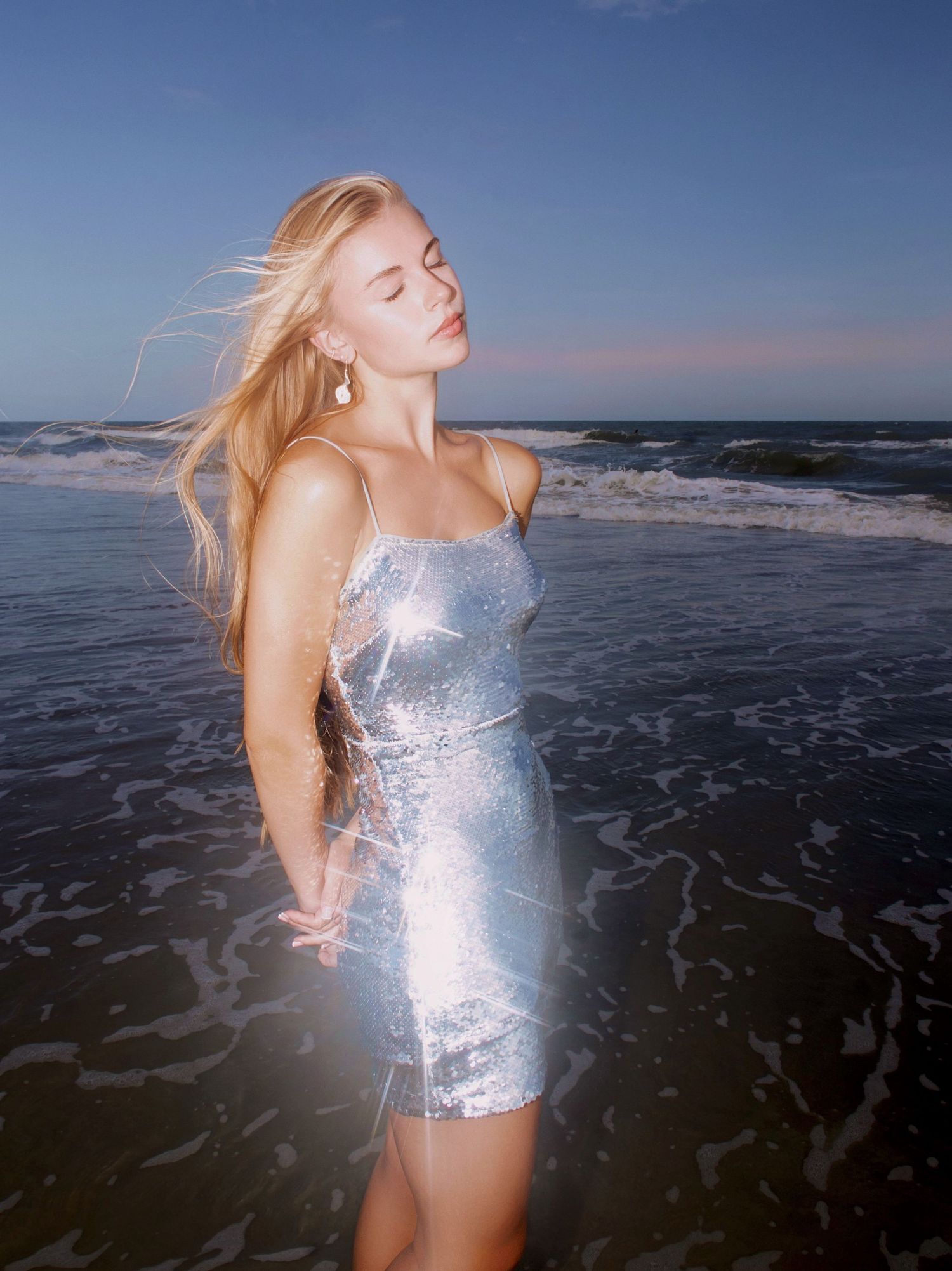 A figure in a shimmering silver mini dress poses on a beach at sunset with waves crashing behind them.