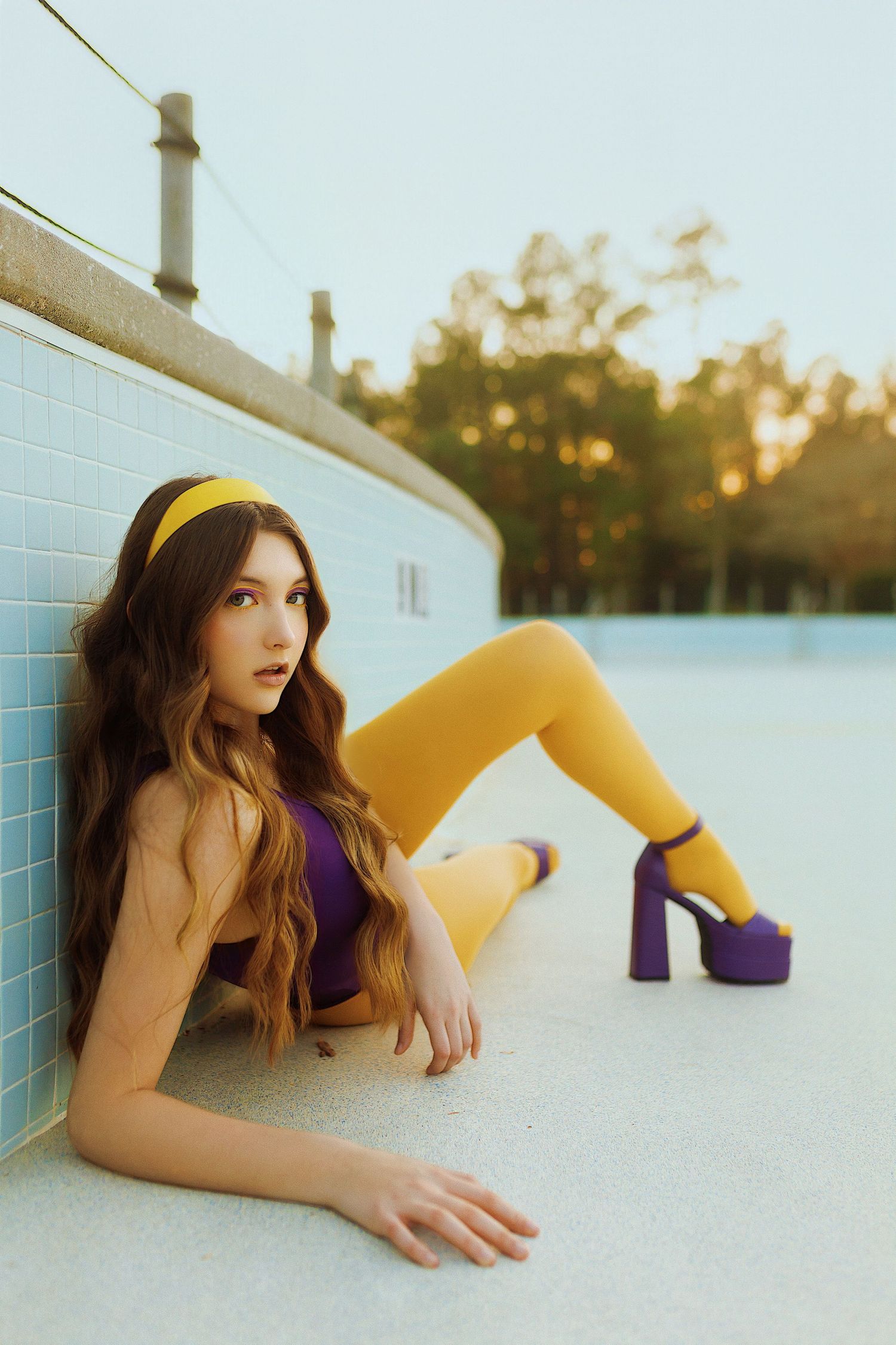 Fashion portrait at sunset with model in purple top and yellow tights reclining against white wall with platform heels.