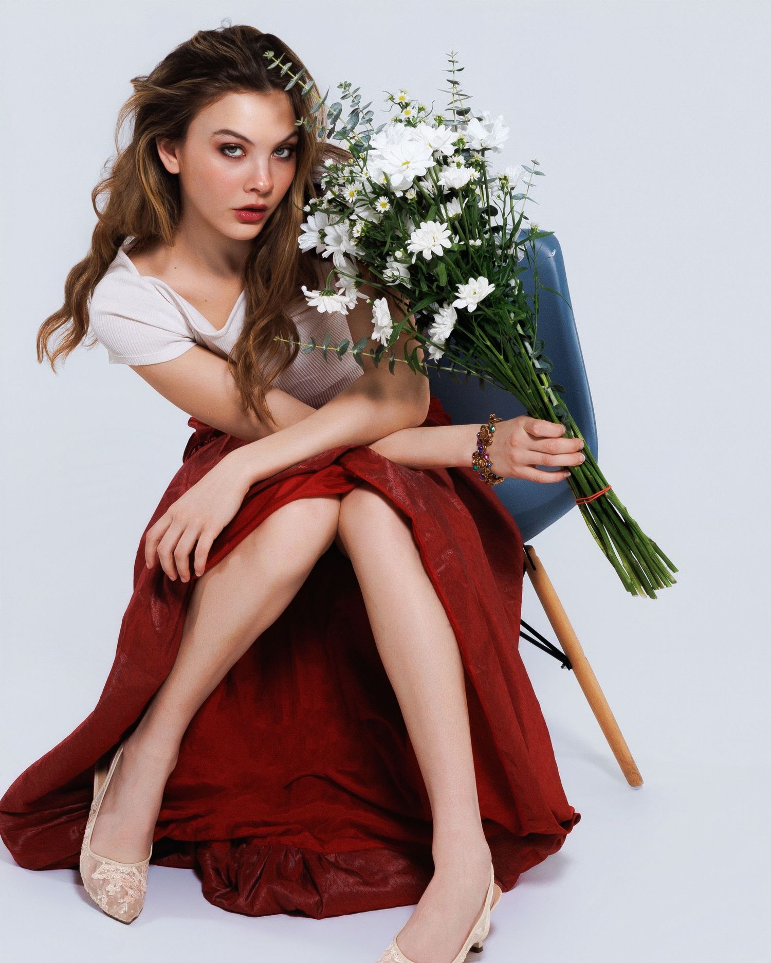 A glamorous model in a red dress poses with a bouquet of white flowers while sitting on a wooden chair against a white backdrop.