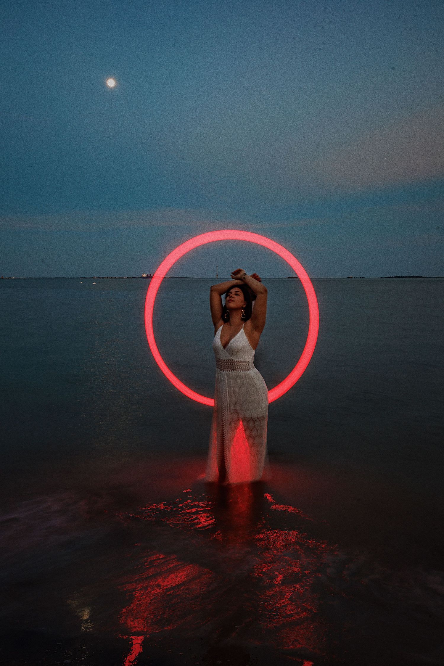 A person stands in shallow ocean water at dusk creating light painting effects with a red LED light ring.