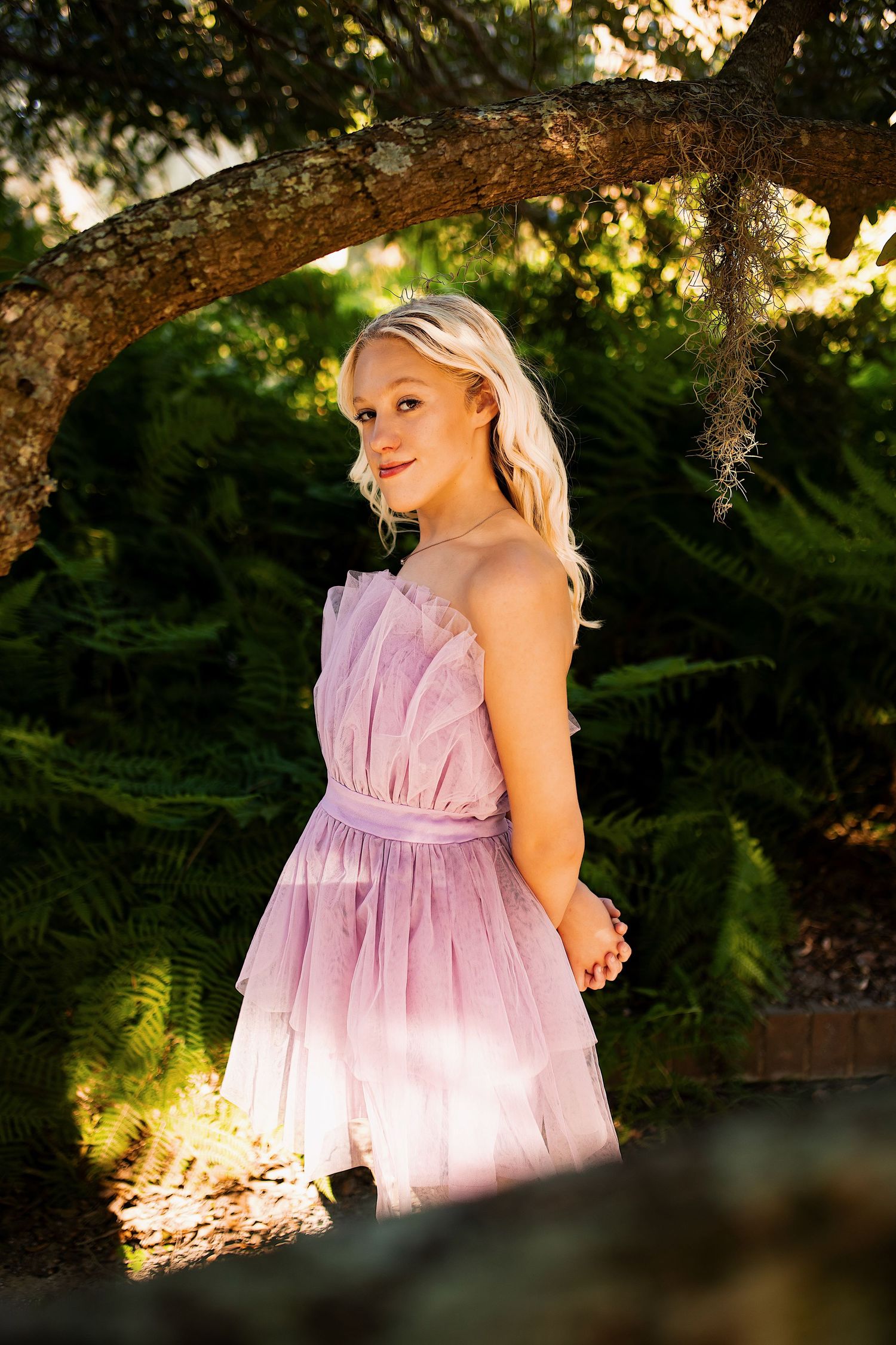 A person in a flowing pink tulle dress poses outdoors in dappled sunlight under trees during a photo session.