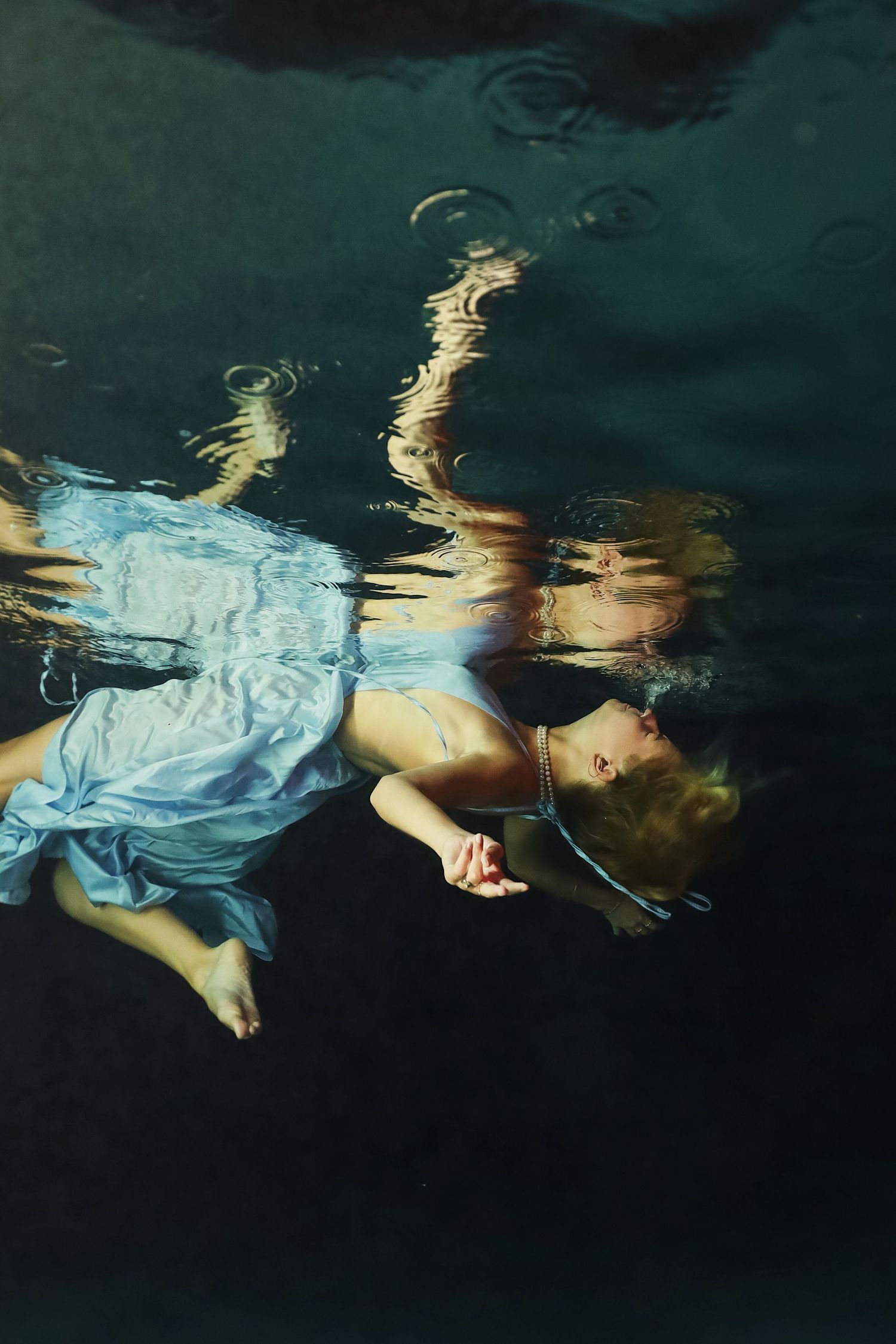 A figure wearing light blue fabric drifts underwater creating an ethereal and dreamlike reflection in dark water.