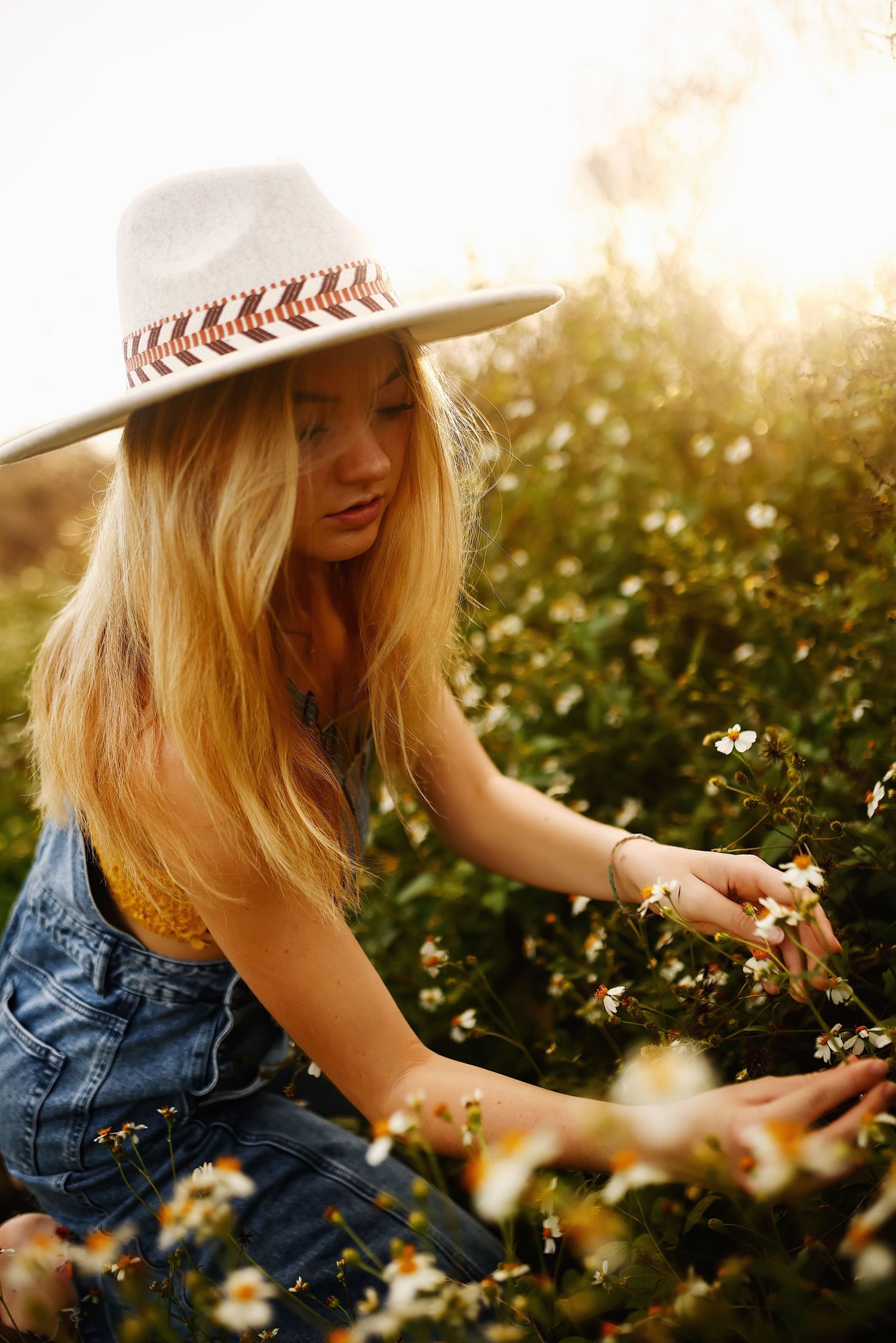 Person in denim and hat reaches for wildflowers in a sunlit meadow during golden hour.