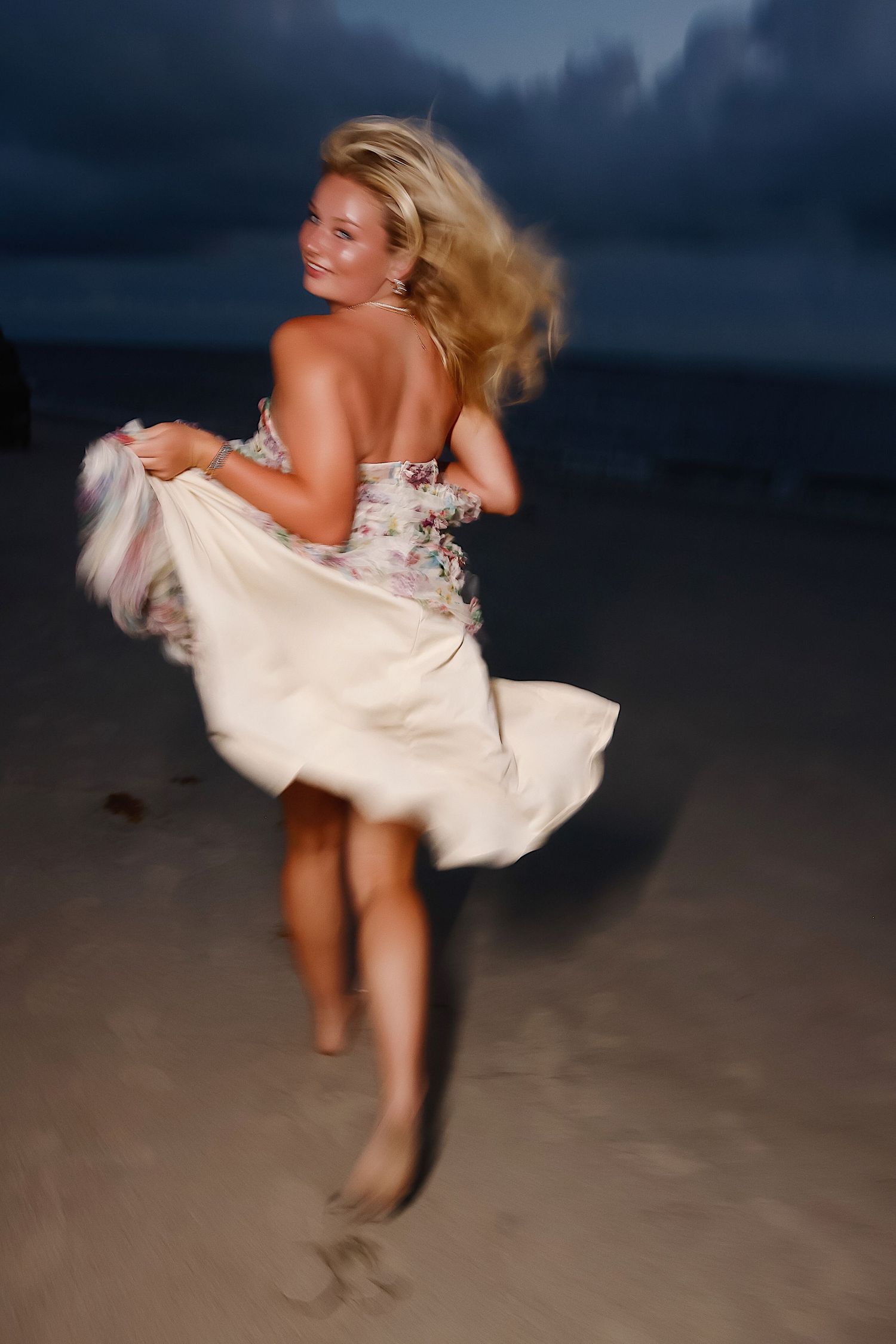 A figure in a white dress twirls on a beach at dusk creating a dynamic motion blur effect.