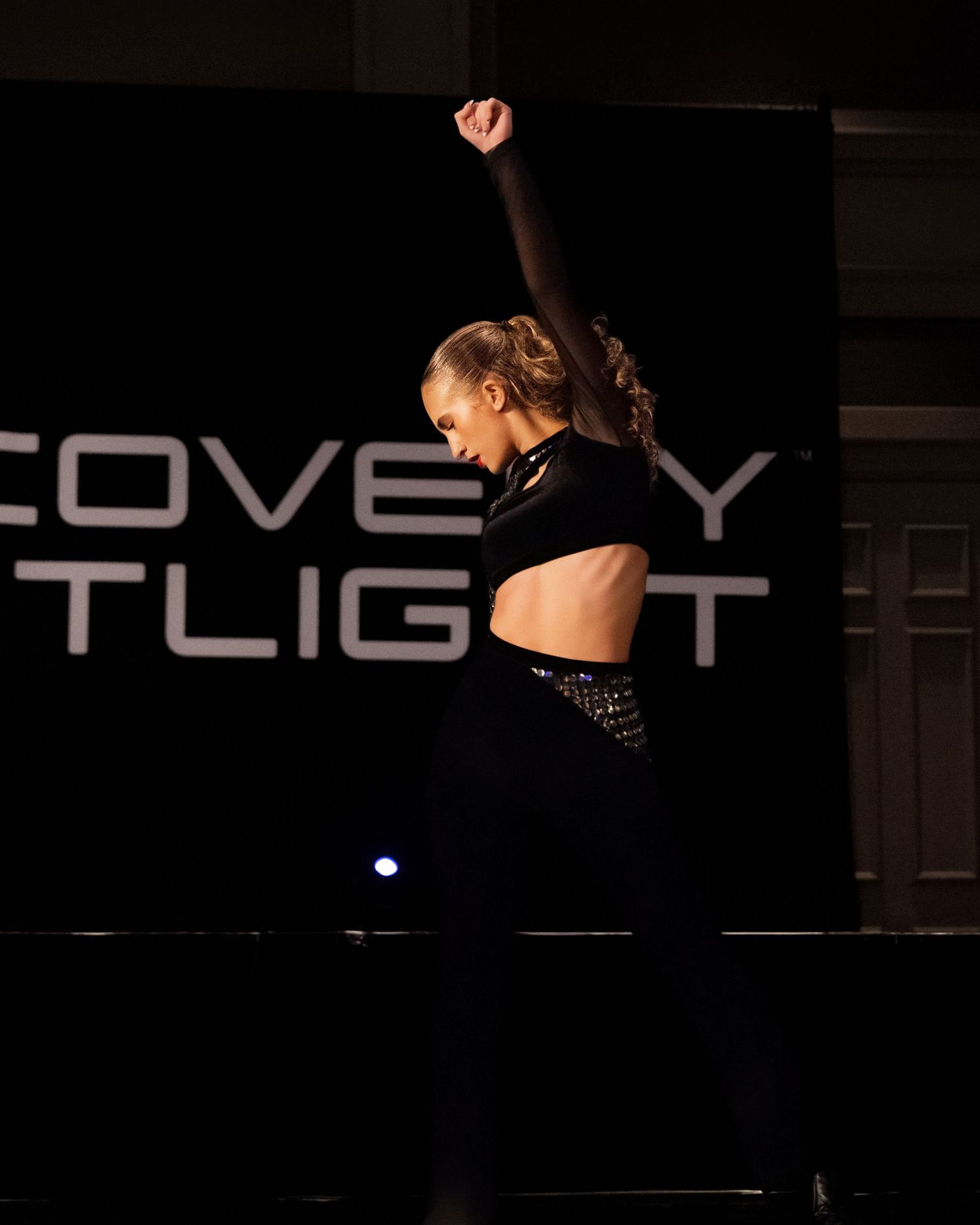 A dancer in black performs a dynamic movement against a dark stage with illuminated text visible in the background.