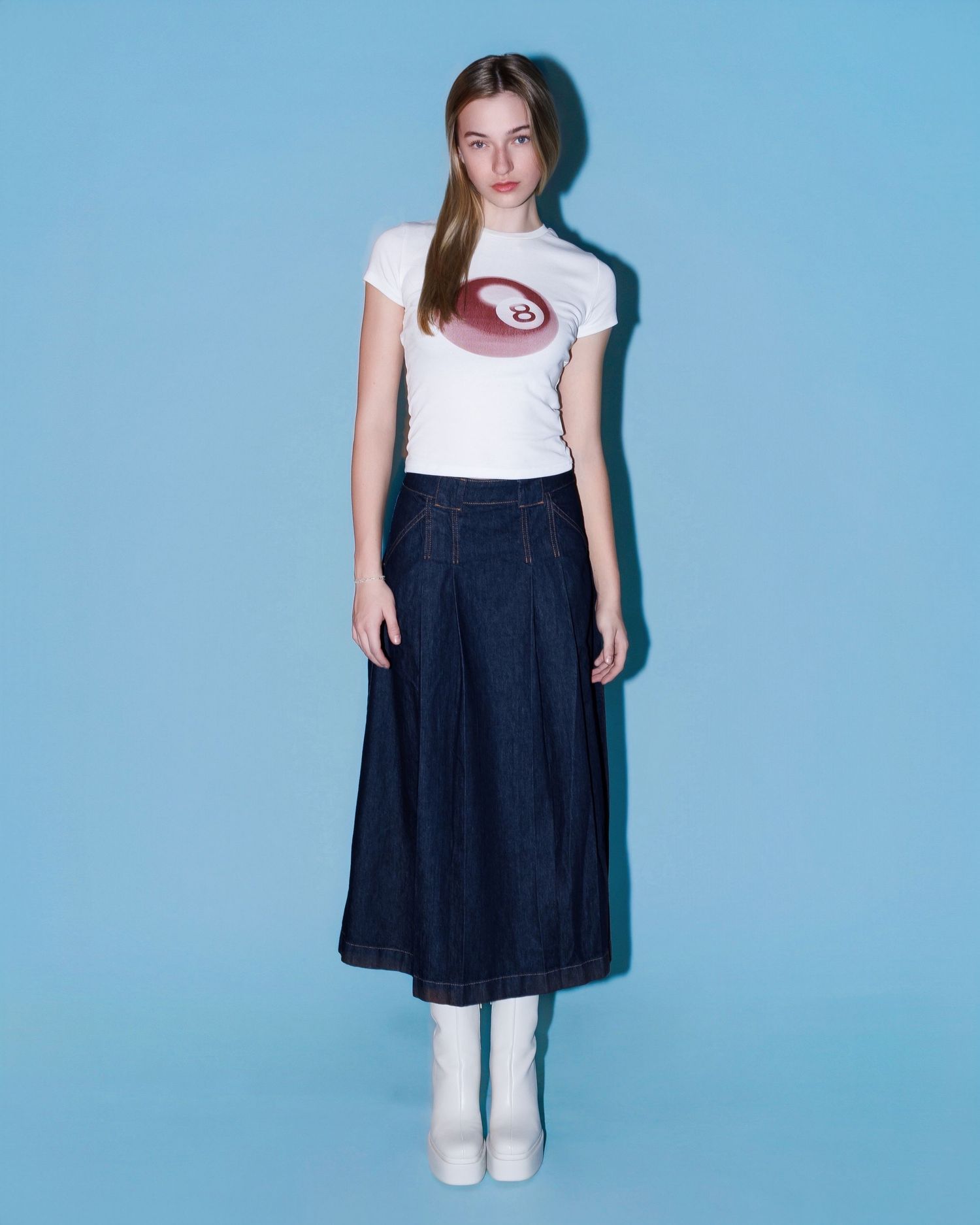 A person wearing a white graphic t-shirt, navy midi skirt and white boots poses against a light blue background.