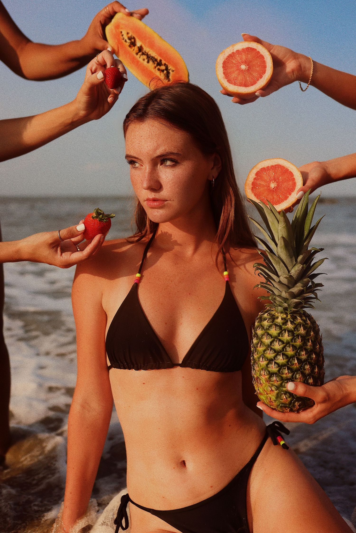 Woman in black bikini at beach surrounded by fresh tropical fruits like pineapple, grapefruit and strawberry.