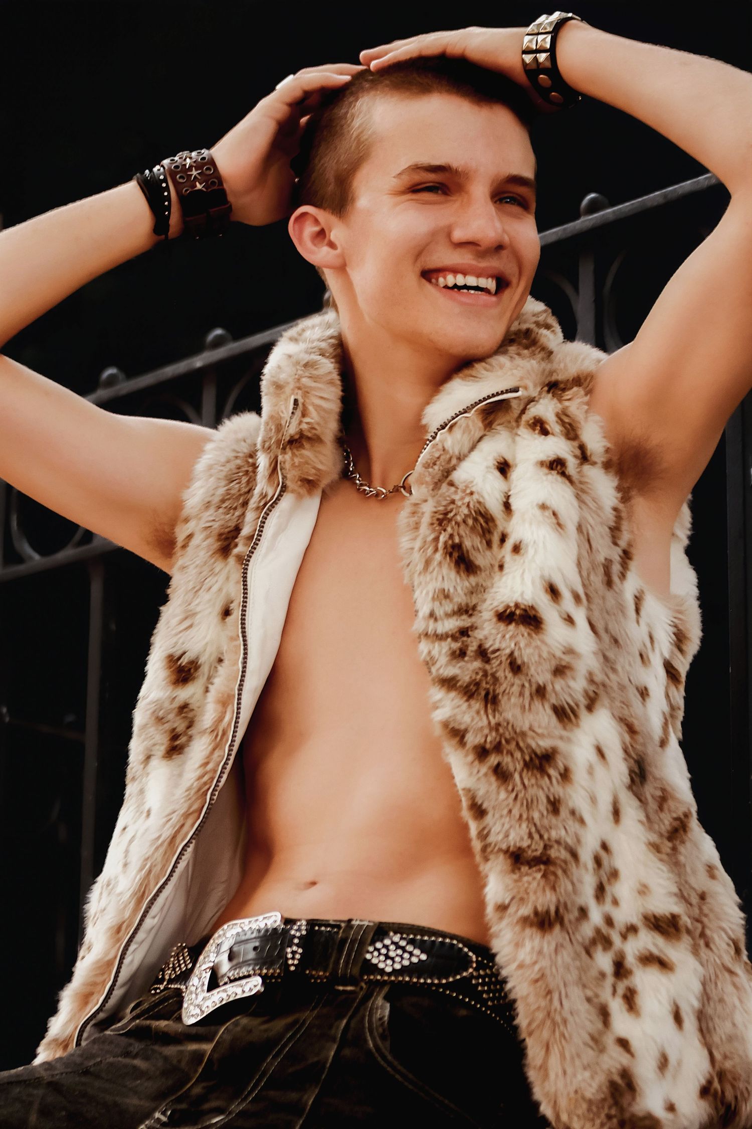 Someone poses joyfully in a furry spotted vest and dark bottoms against a black backdrop.