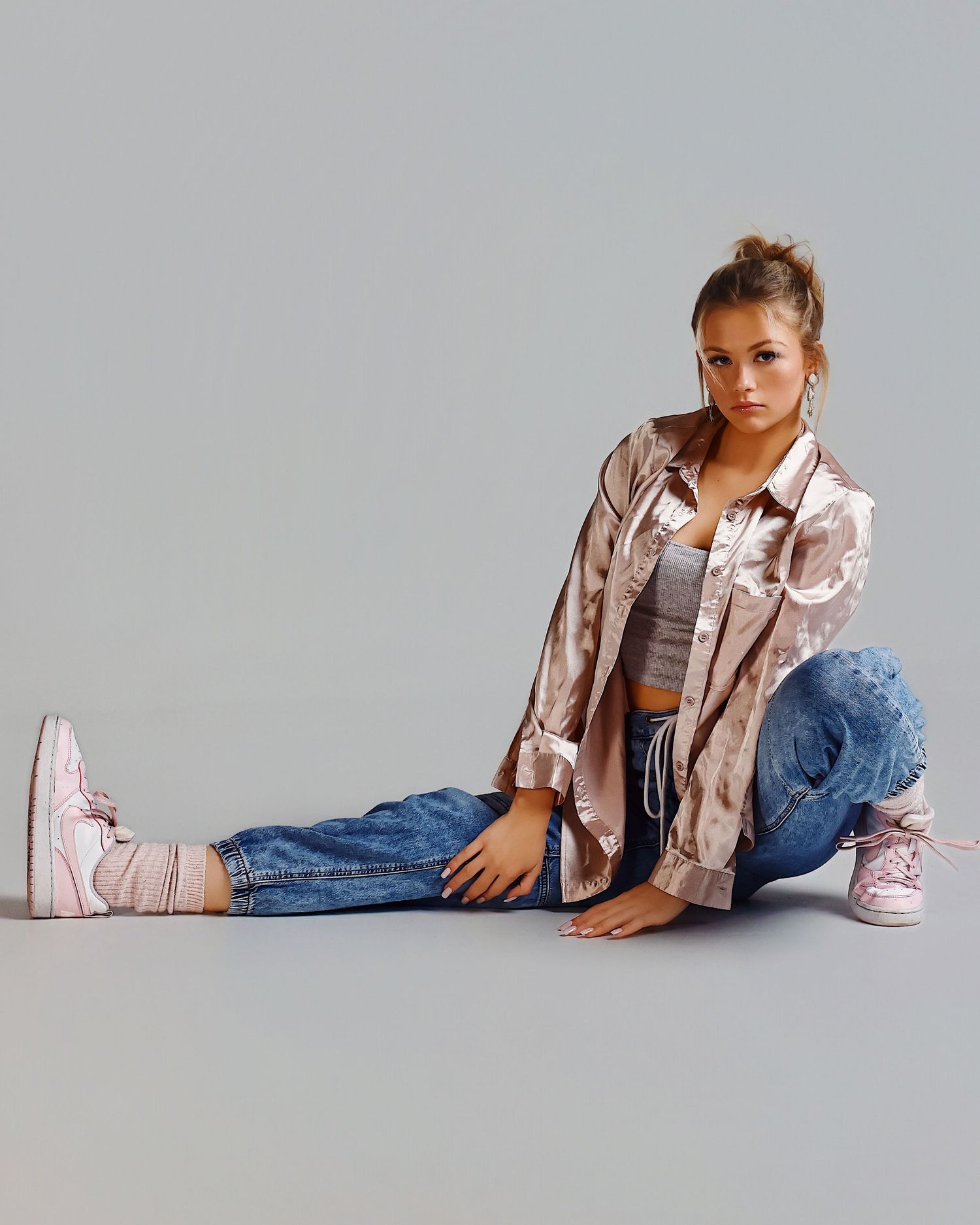 Model in casual attire sitting on floor wearing pink satin jacket, denim jeans and pink sneakers against grey background.