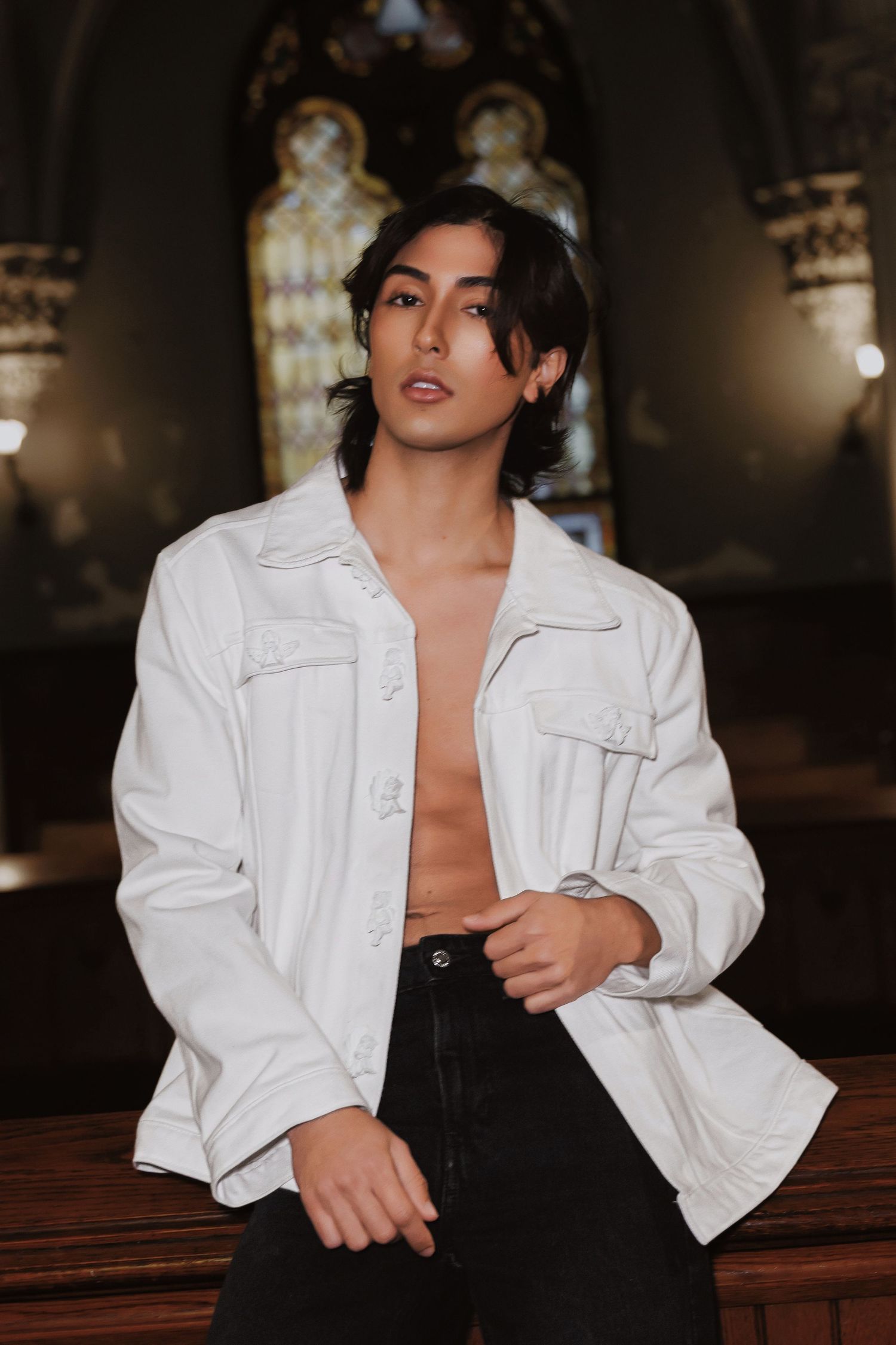 Stylish model in white open jacket and black pants poses dramatically in gothic church interior with stained glass windows.