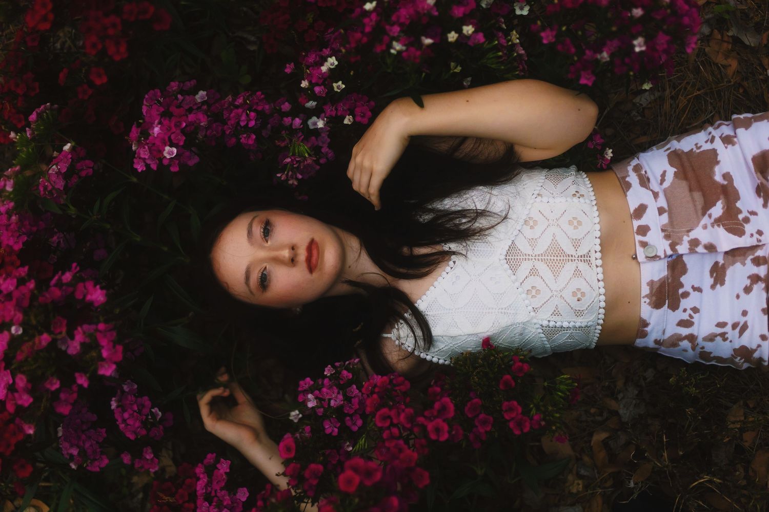 Person in white lace dress lying on dark ground surrounded by scattered pink flower petals in romantic setting.