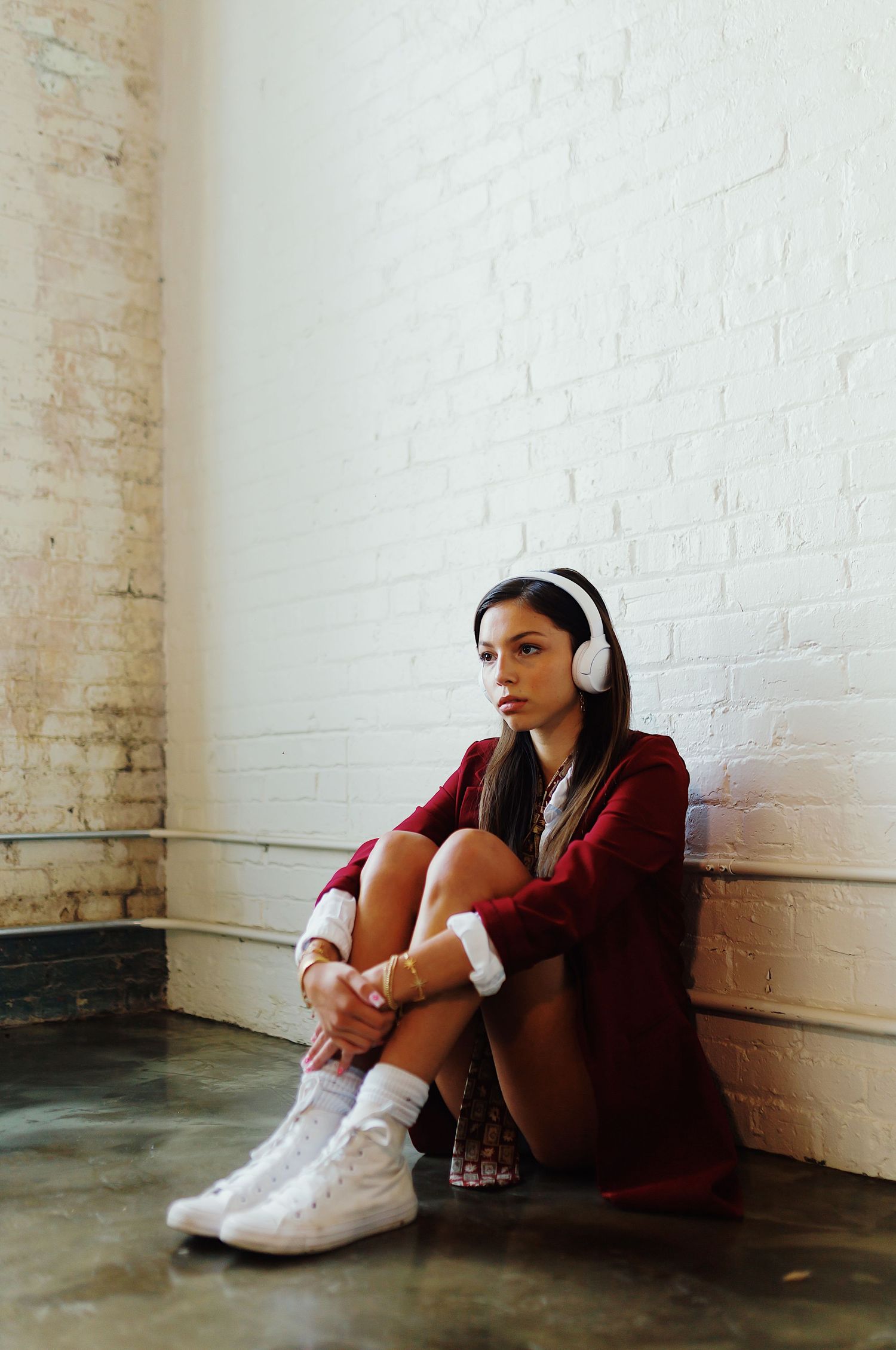 A person wearing headphones and burgundy clothing sits alone against a white brick wall in a minimalist setting.