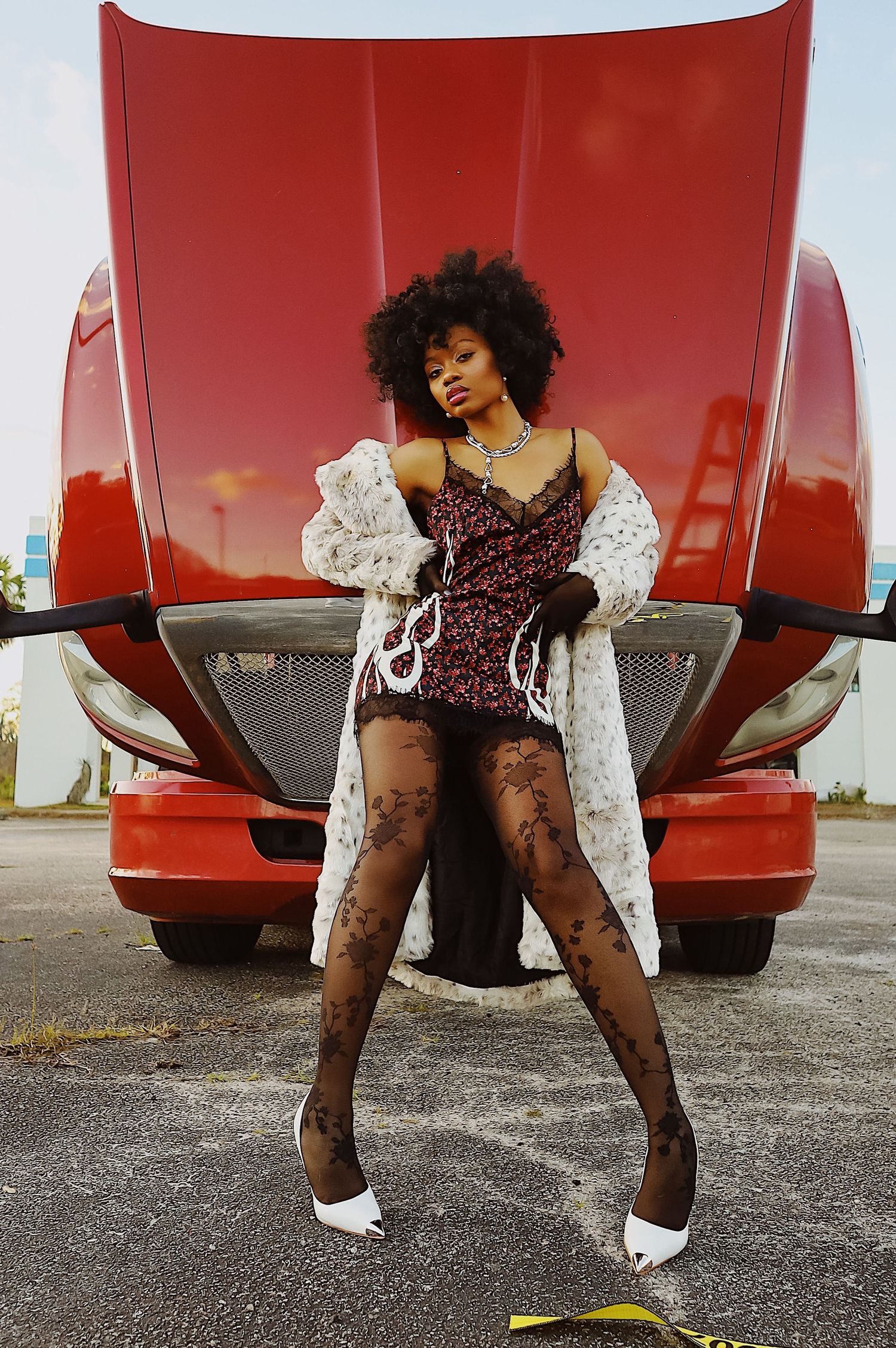 A person in patterned stockings and white boots poses playfully in front of a red vehicle.