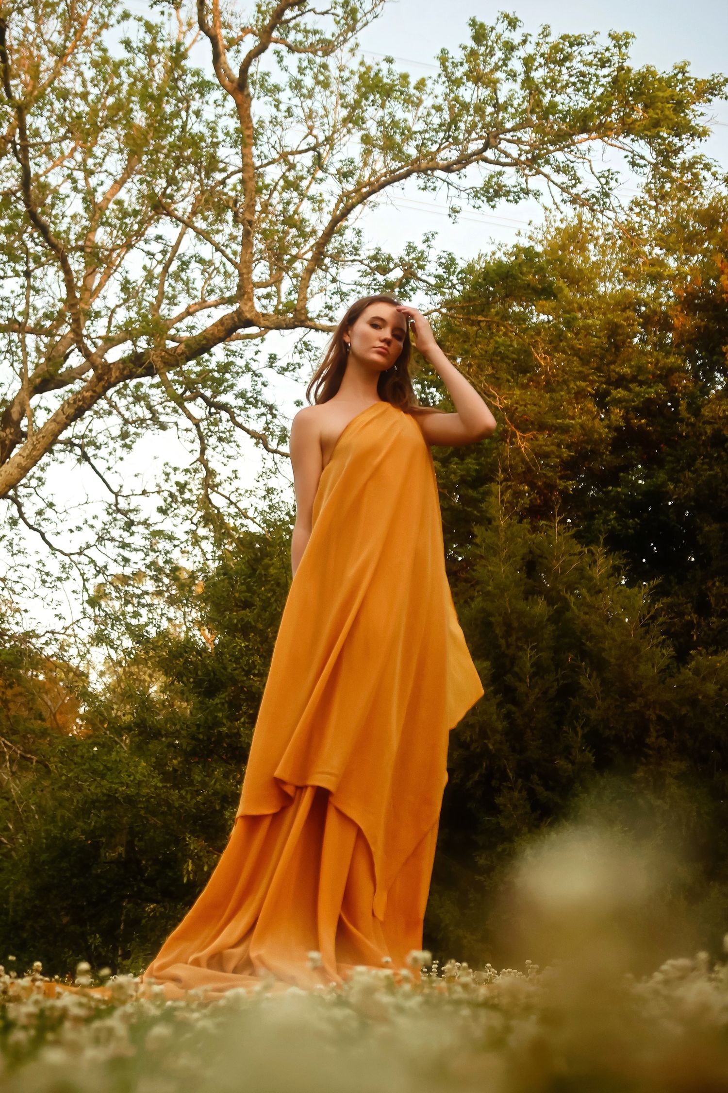 Elegant marigold-colored flowing dress photographed in a dreamy natural setting with trees at golden hour.