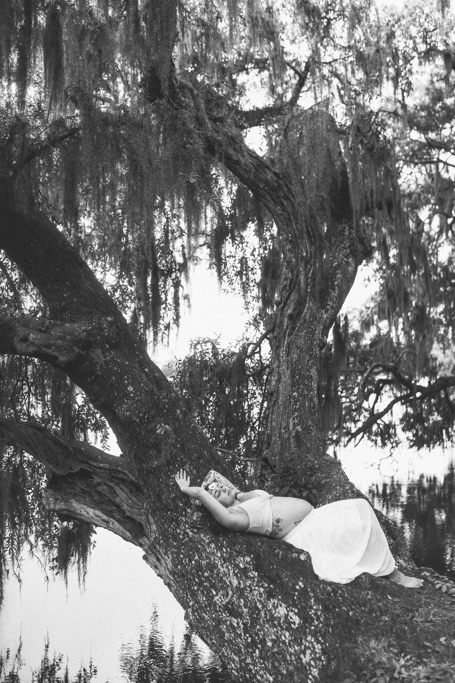 Black and white photo of a woman in a white dress reclining on a large tree branch draped with Spanish moss.