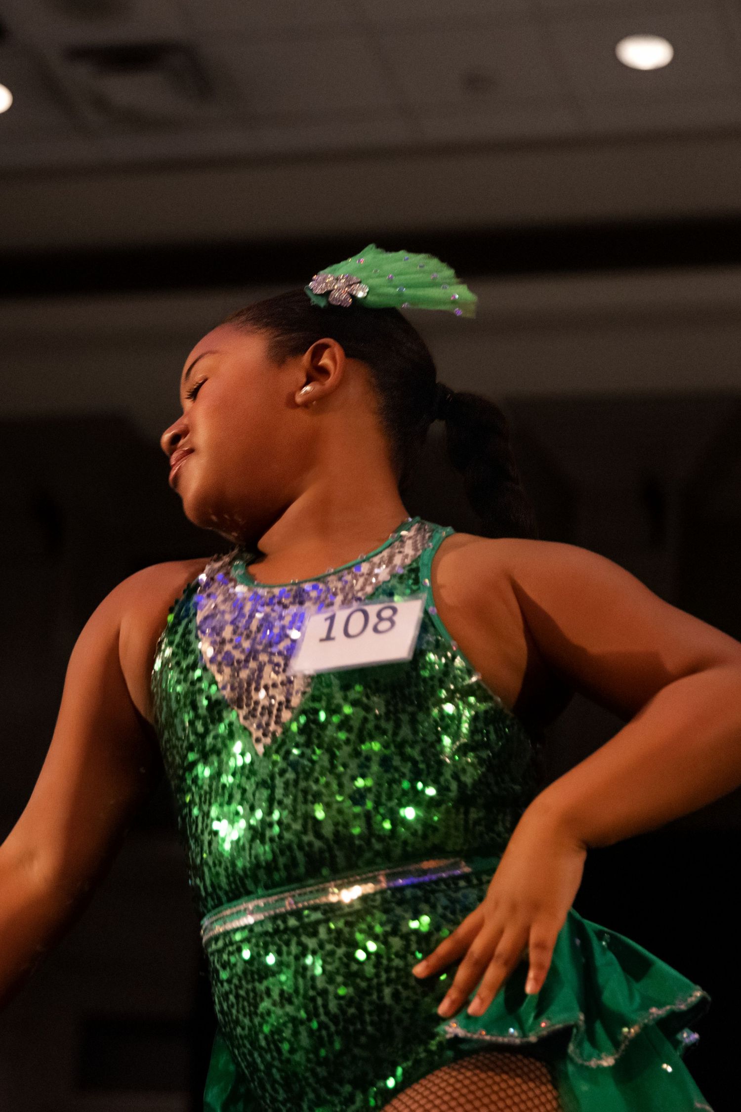 Dancer in sparkly green costume performs graceful movements during a performance on a dark stage.
