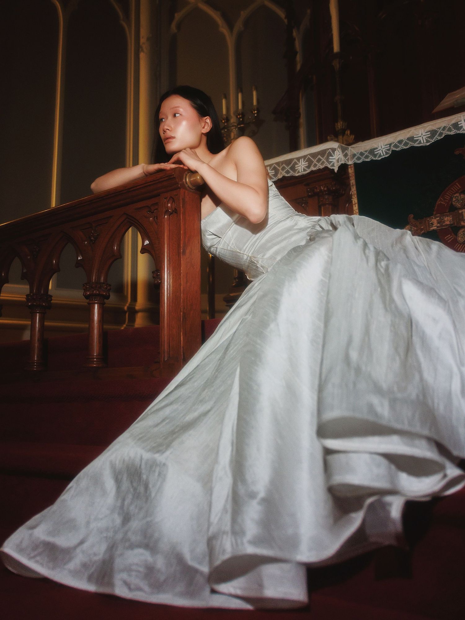 A person in an elegant white wedding gown rests gracefully against ornate wooden church pews in dramatic lighting.