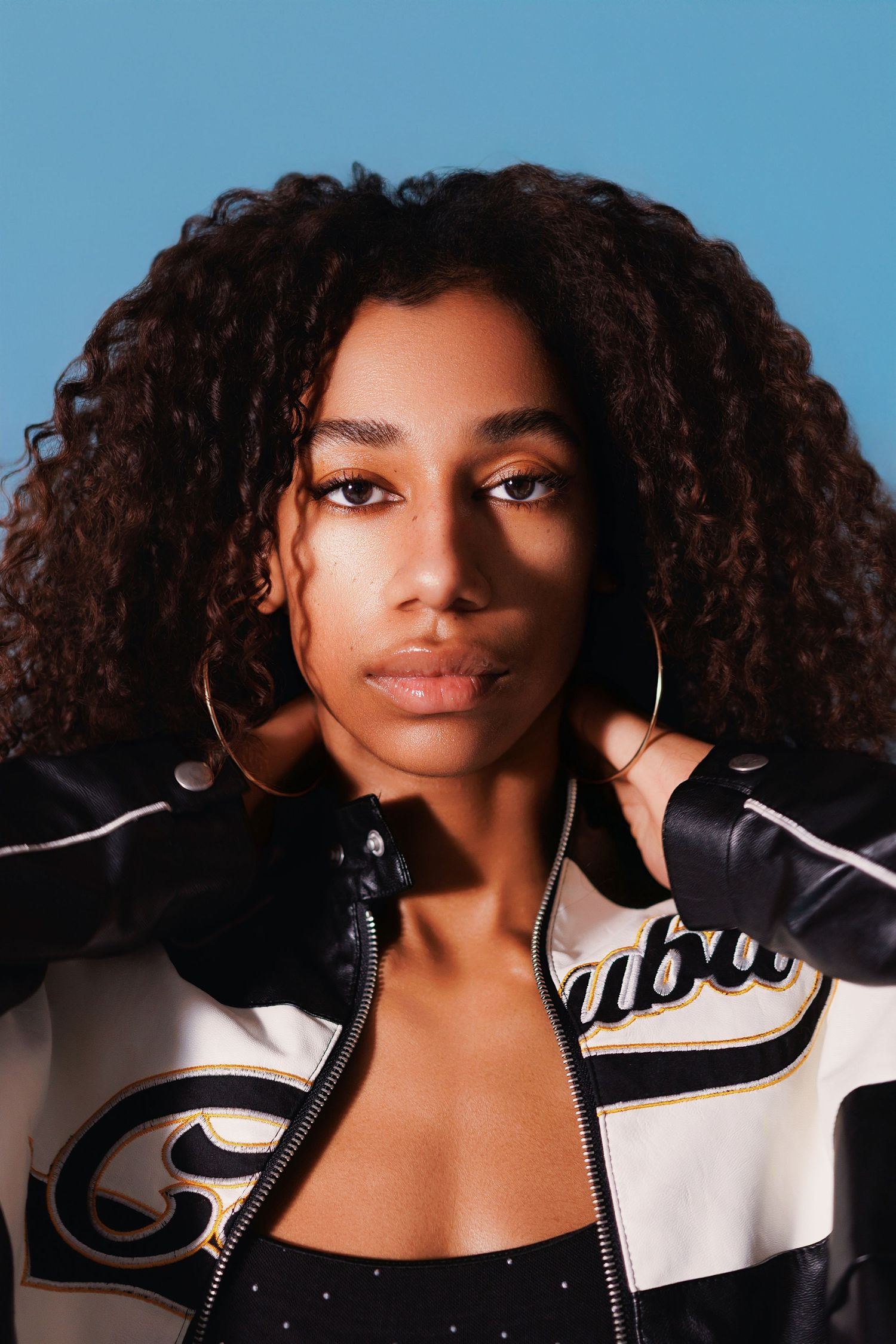 Portrait of a person wearing a stylish black and white leather jacket with curly hair and hoop earrings against blue backdrop.