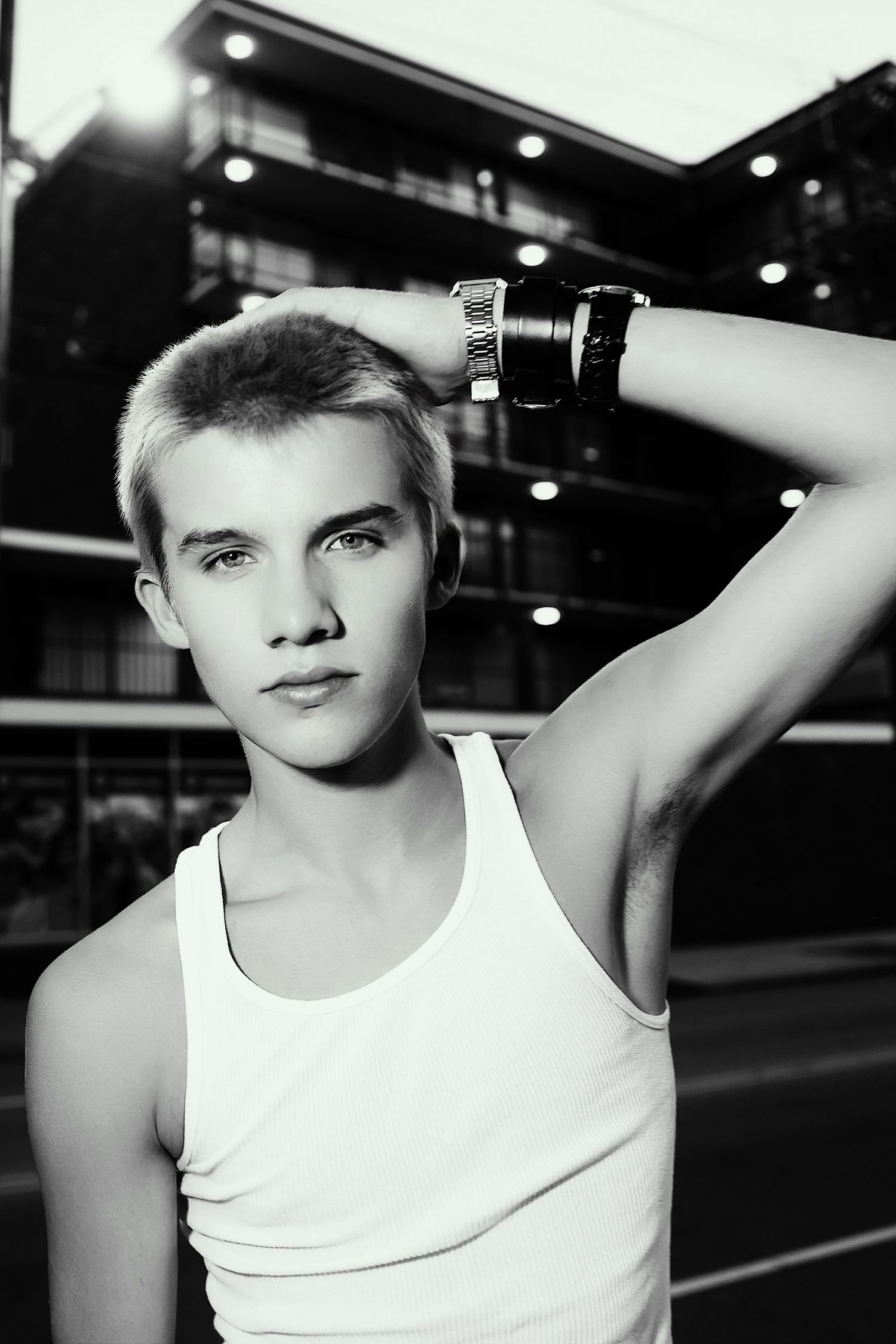 Black and white portrait of a figure in a white tank top wearing a watch, with dramatic urban lighting.