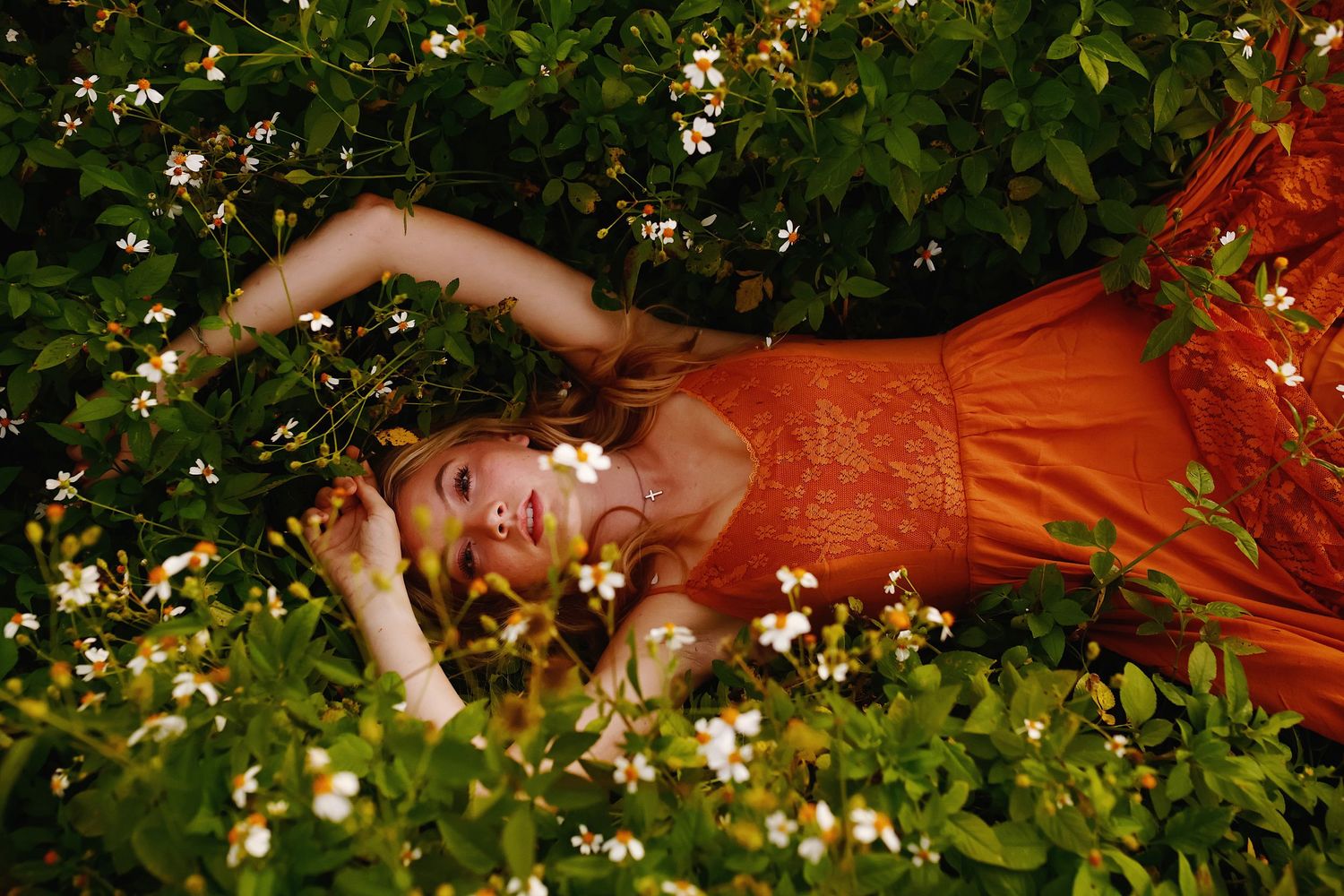 A person in a flowing red dress lies gracefully among lush green foliage and white flowers in an artistic outdoor photo.
