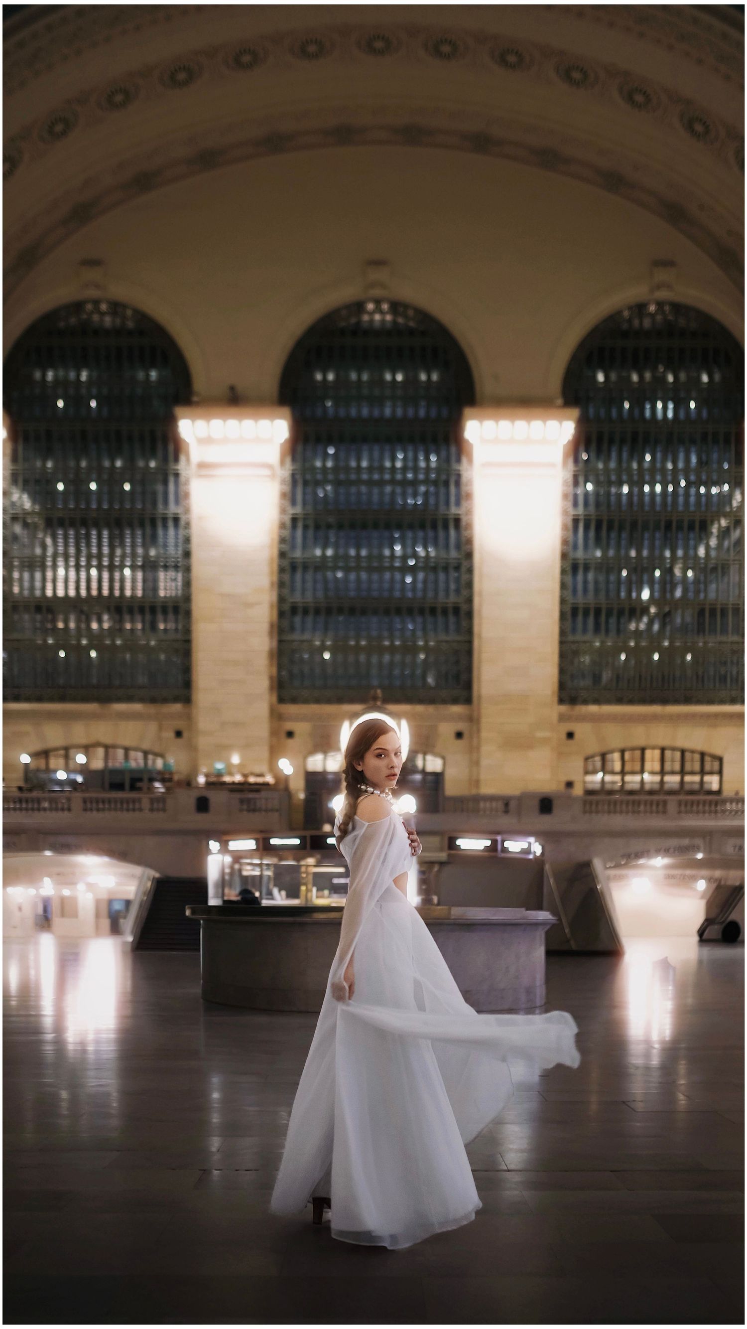 A figure in a flowing white dress twirls and dances gracefully inside the grand hall of Grand Central Terminal in New York City.