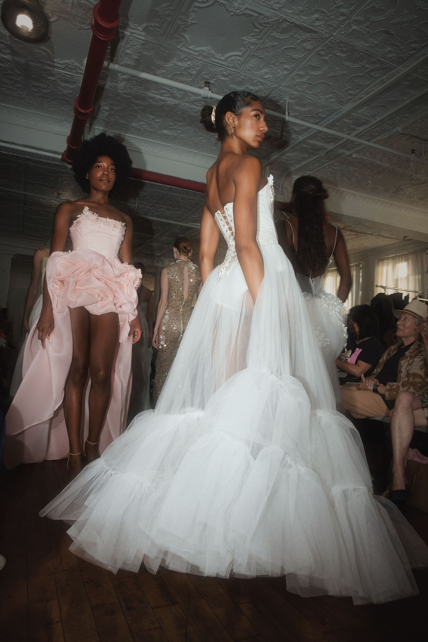Models showcase flowing white tulle wedding gowns and pink dresses on a fashion runway during a bridal show.
