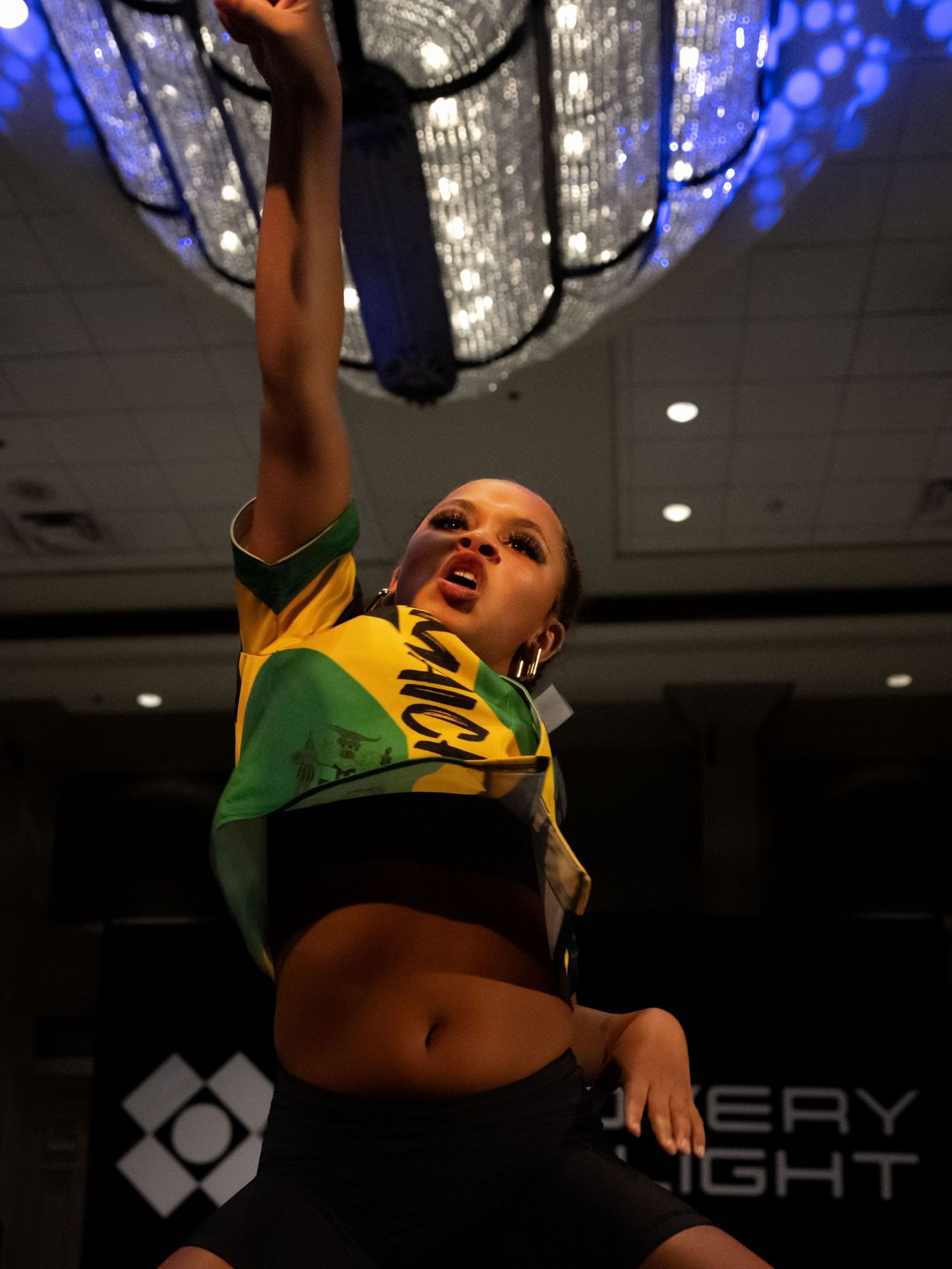 A dancer in yellow and green performs an energetic dance move against a dark background with blue lighting effects.