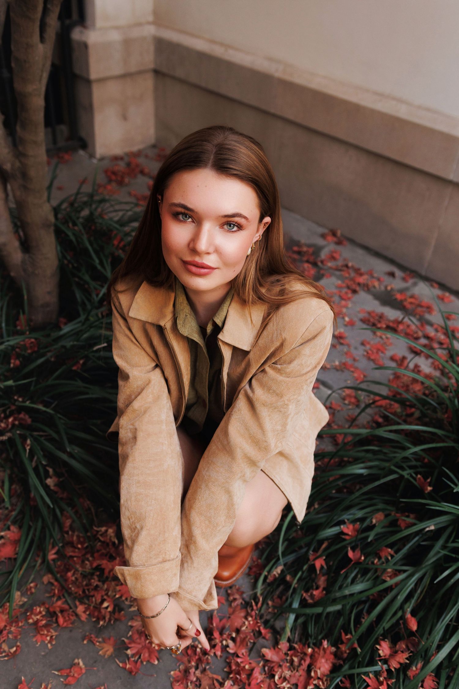A person wearing a beige trench coat crouches next to red autumn leaves and green plants against a light-colored wall.