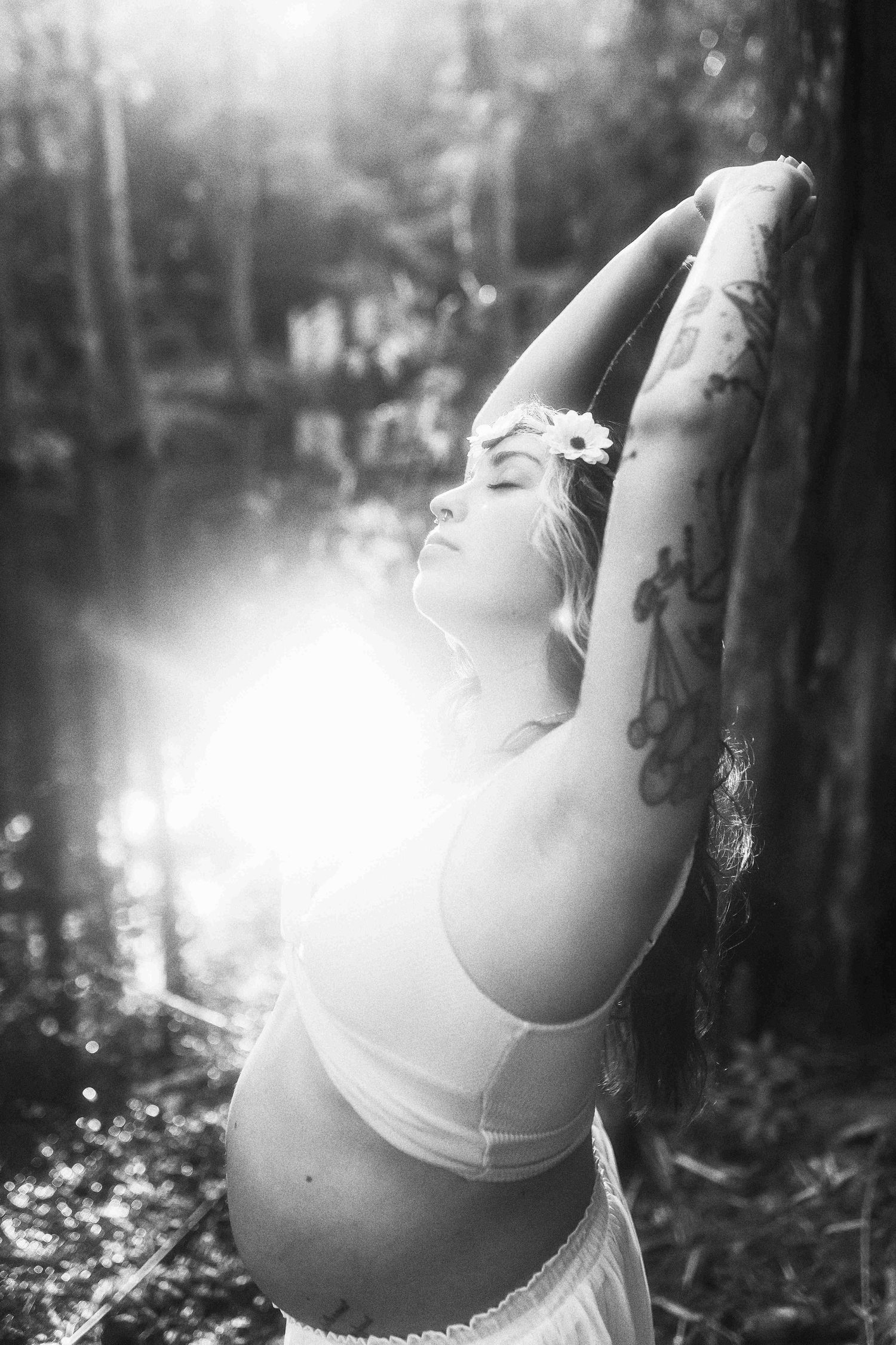 Black and white maternity photo of tattooed woman with flower crown, arms raised, bathed in sunlight near a forest pond.