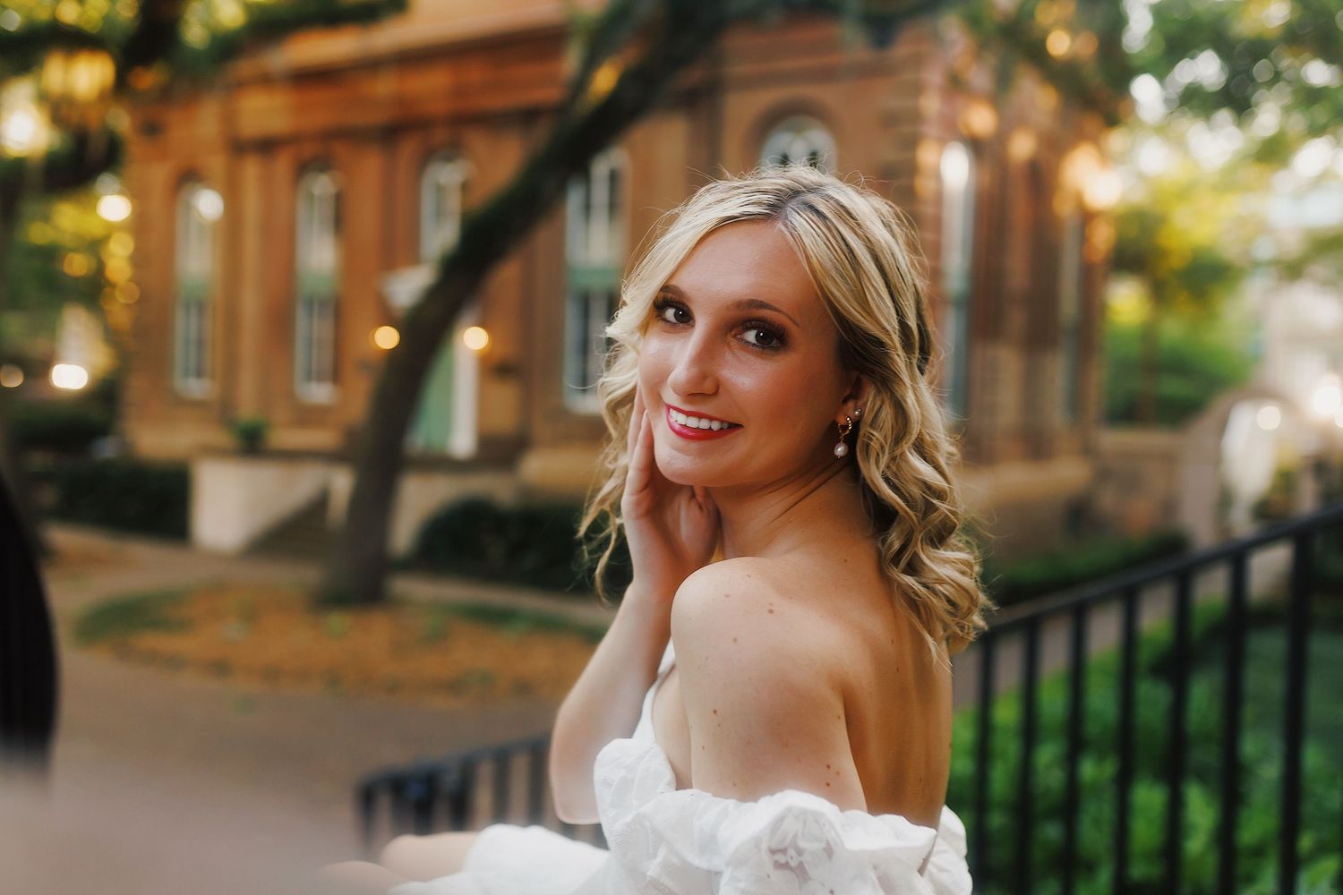 A bride in an off-shoulder white wedding gown smiles while standing outdoors near a brick building.