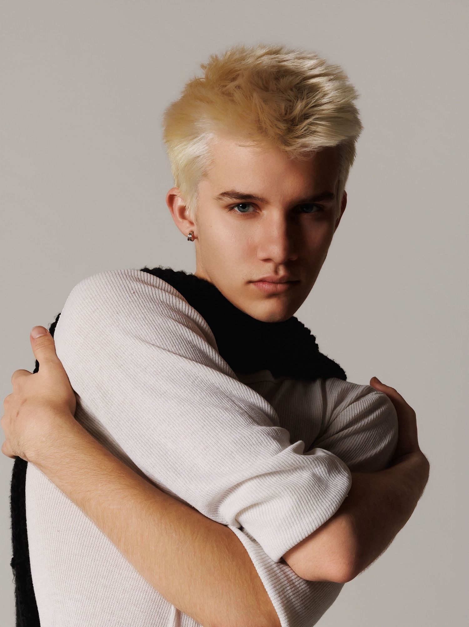 Young person with platinum blonde hair poses in black and white knitwear against neutral backdrop in stylish portrait.