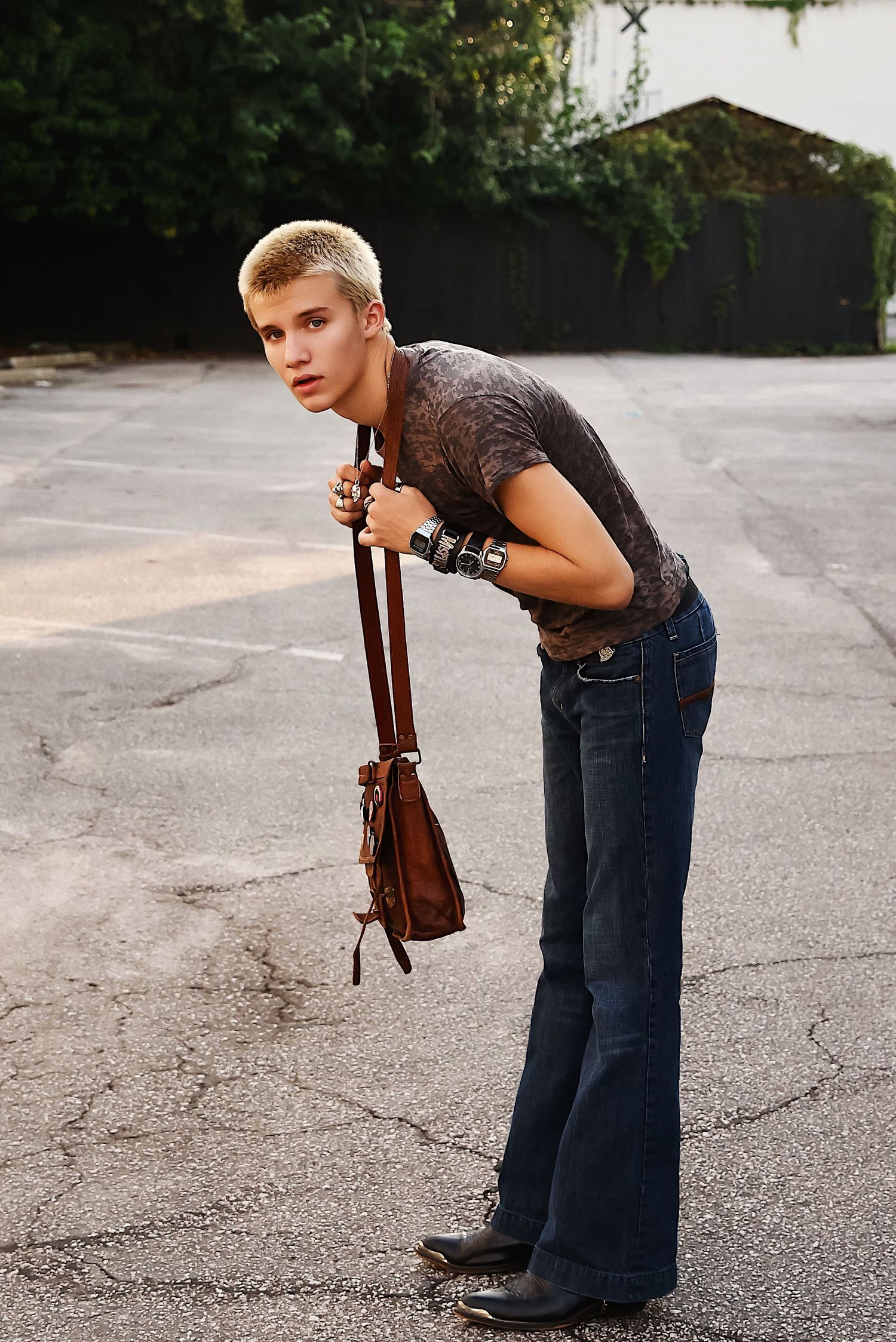 Person in blue jeans and dark t-shirt poses outdoors with a brown leather bag on a paved surface.