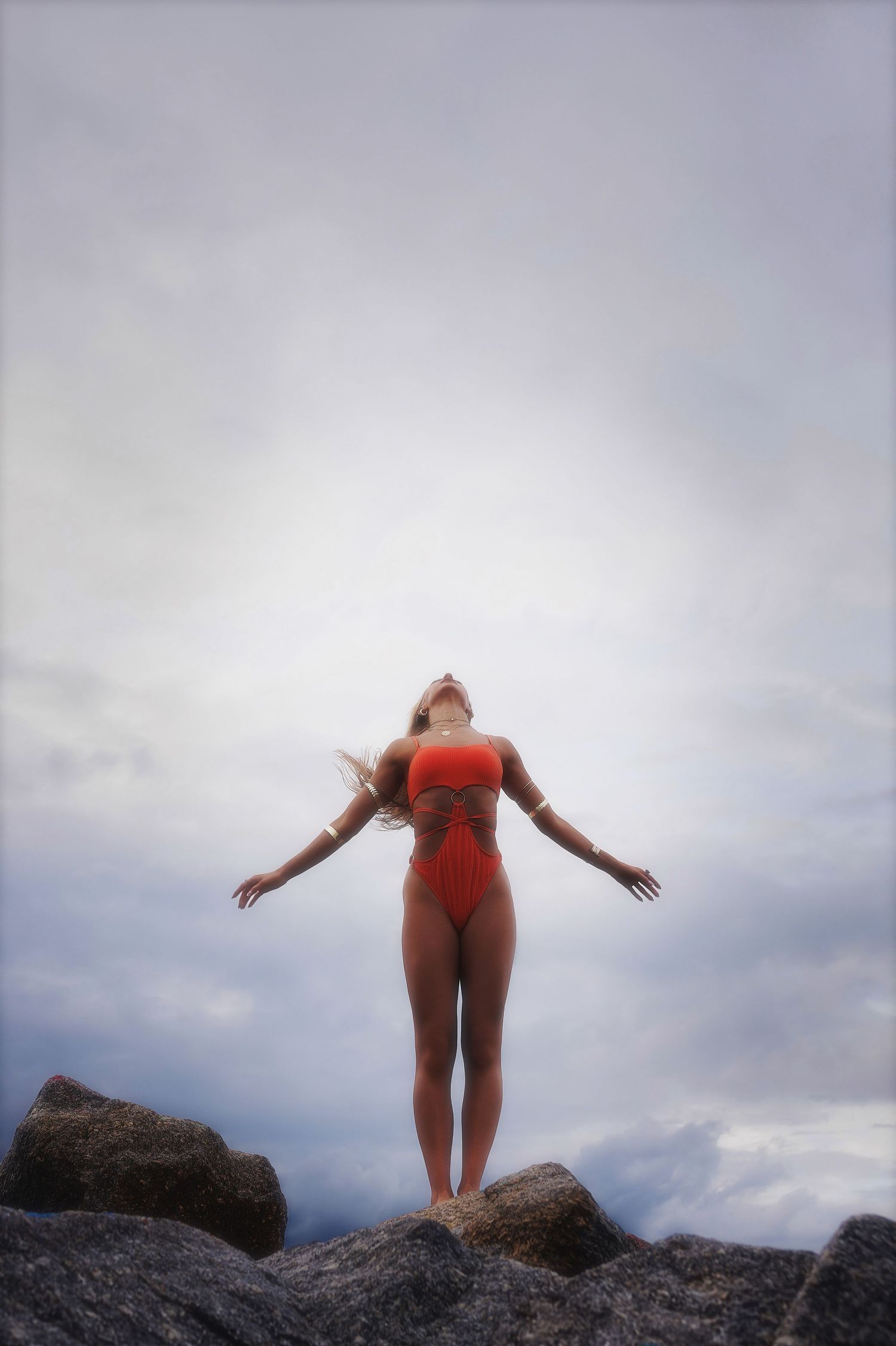 A person in red swimwear stands with arms outstretched on rocky coastal cliffs against a cloudy sky.