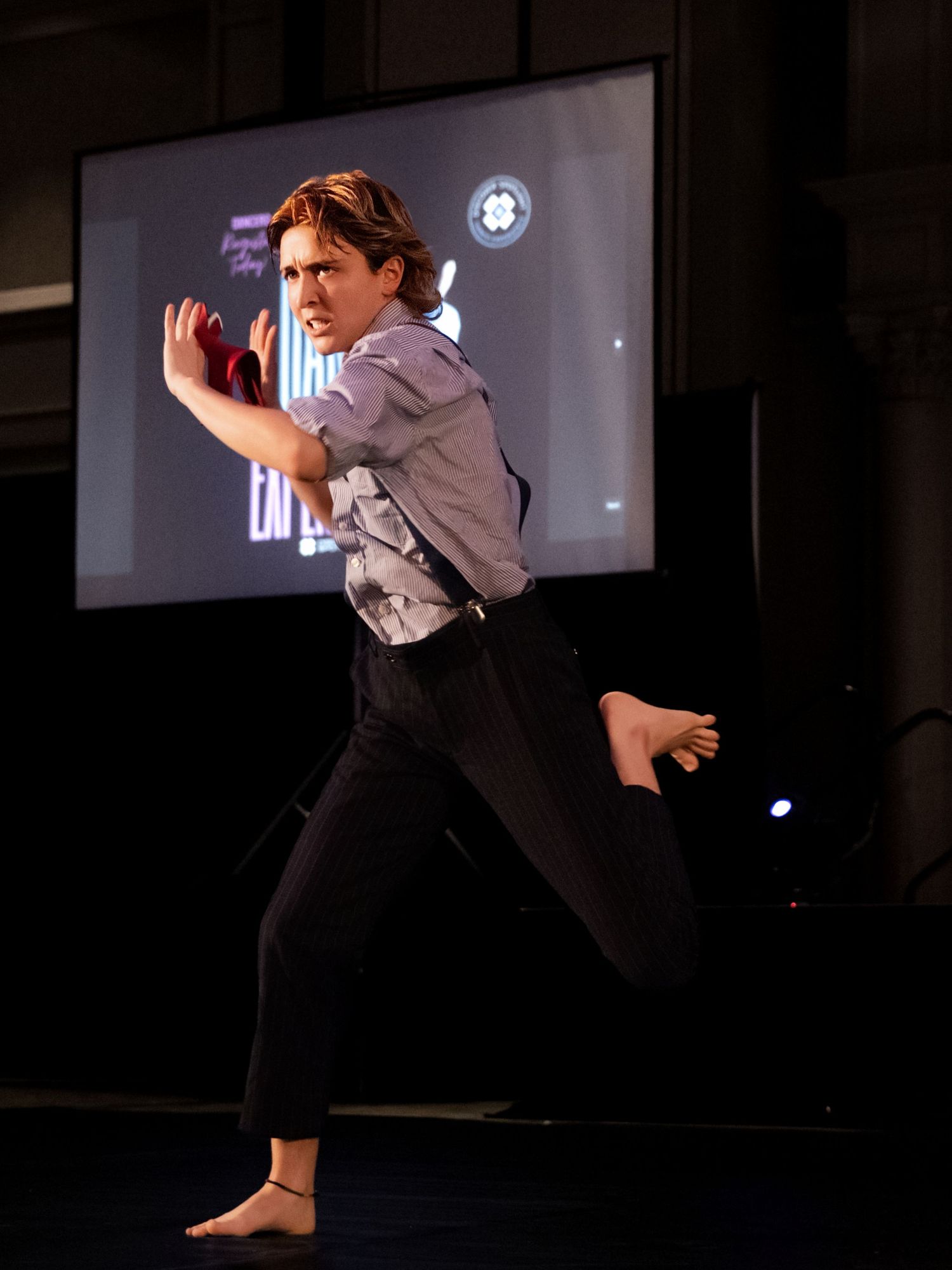 A dancer in black pants and white shirt performs a dynamic leaping movement sequence on a dark stage.