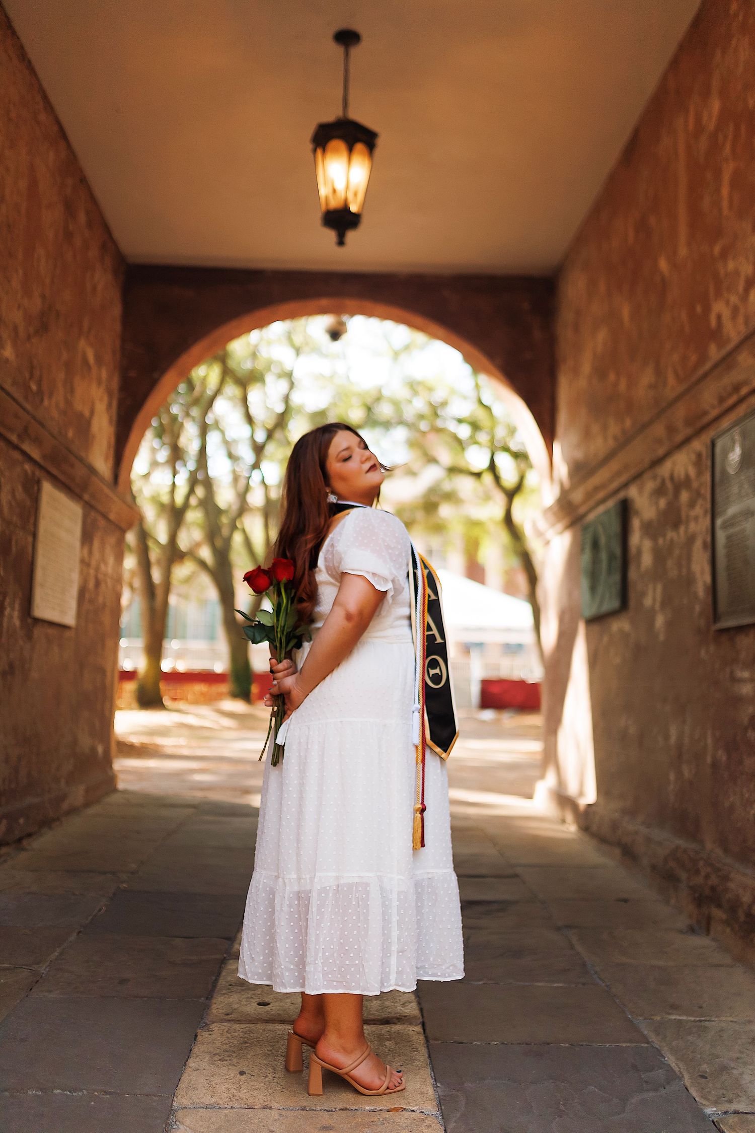 A person in a white eyelet dress stands in a stone archway corridor with dramatic lighting and shadows.