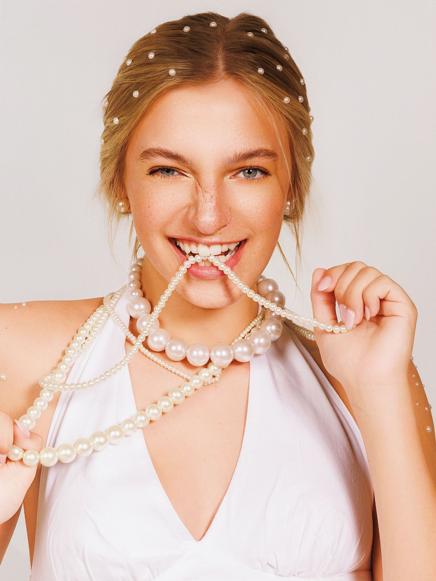 Person in white v-neck top wearing pearl necklaces and hair accessories playfully biting pearl strand against light background.