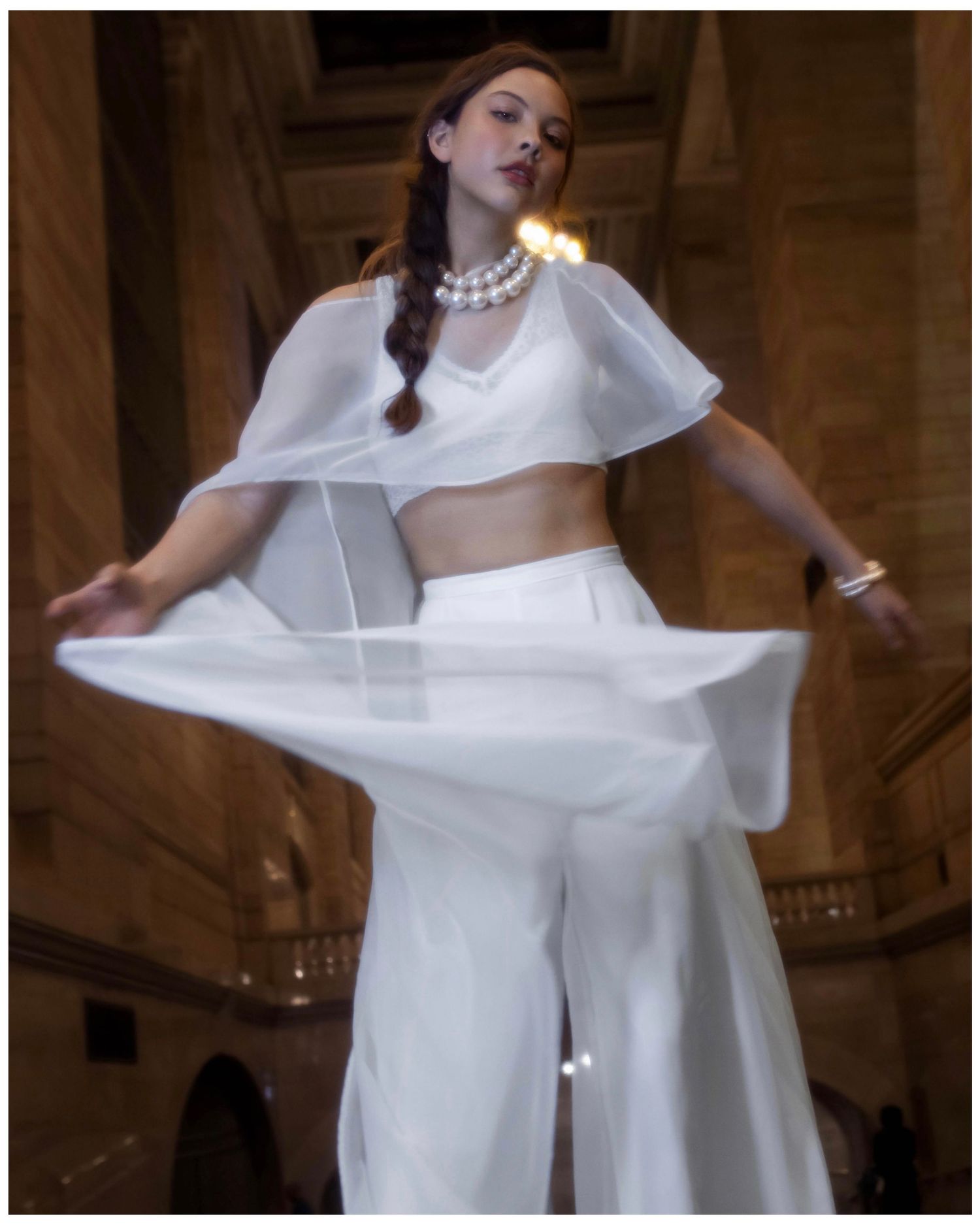 Fashion model wearing an all-white modern ensemble with crop top, wide-leg pants and pearl necklace in architectural setting.