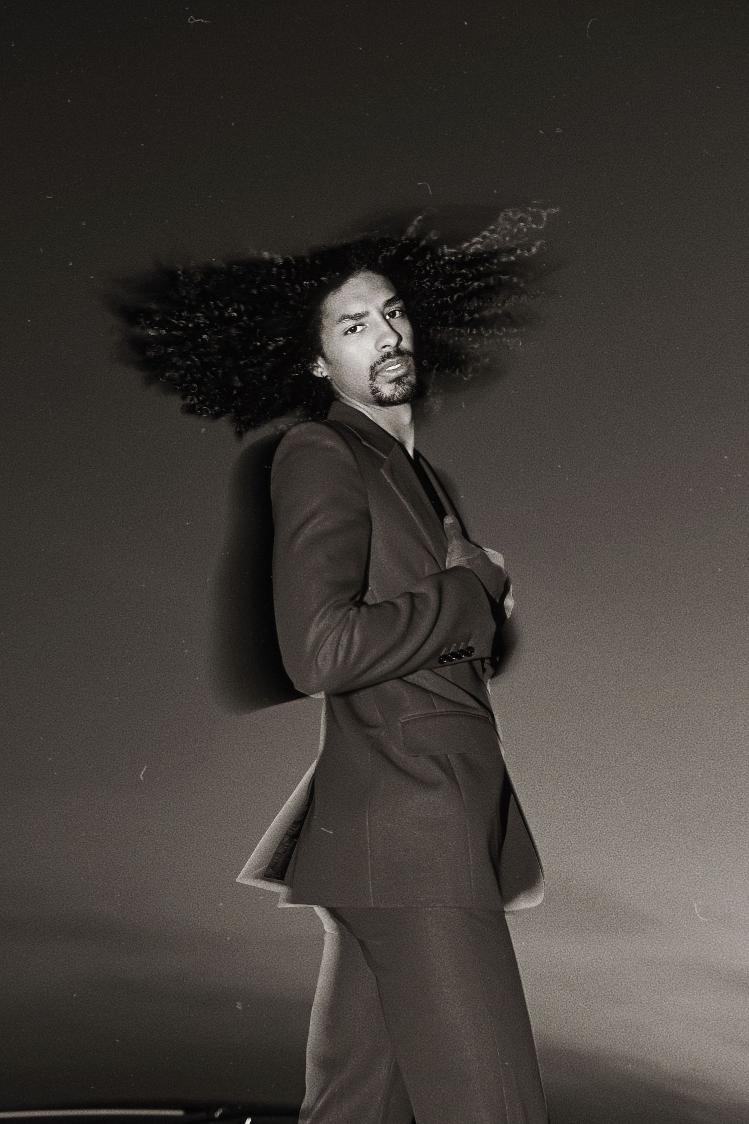 Dynamic black and white photo sequence showing person with dark curly hair spinning in a suit jacket against gray background.
