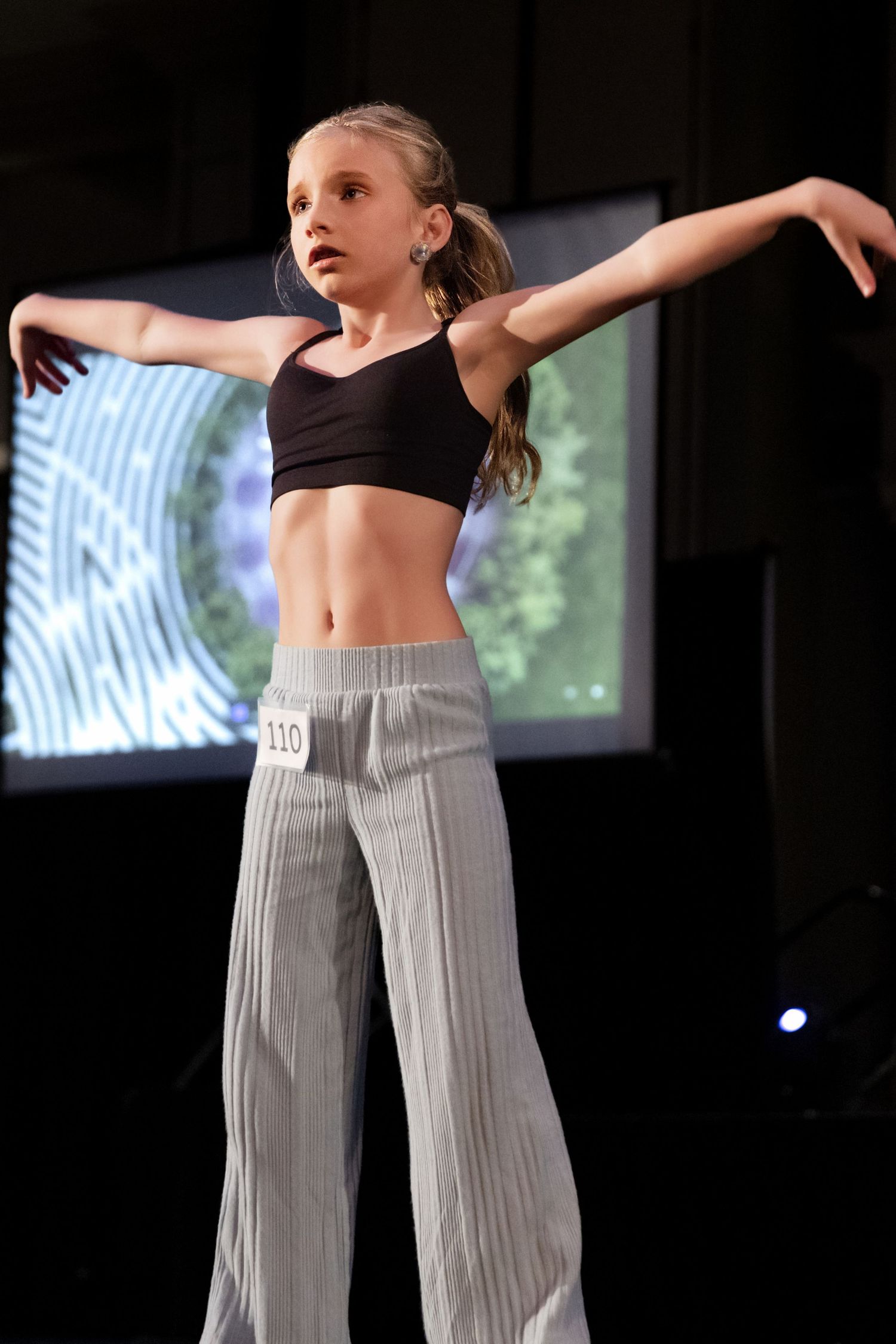 Young dancer in black crop top and gray pants strikes graceful pose during performance on dark stage.