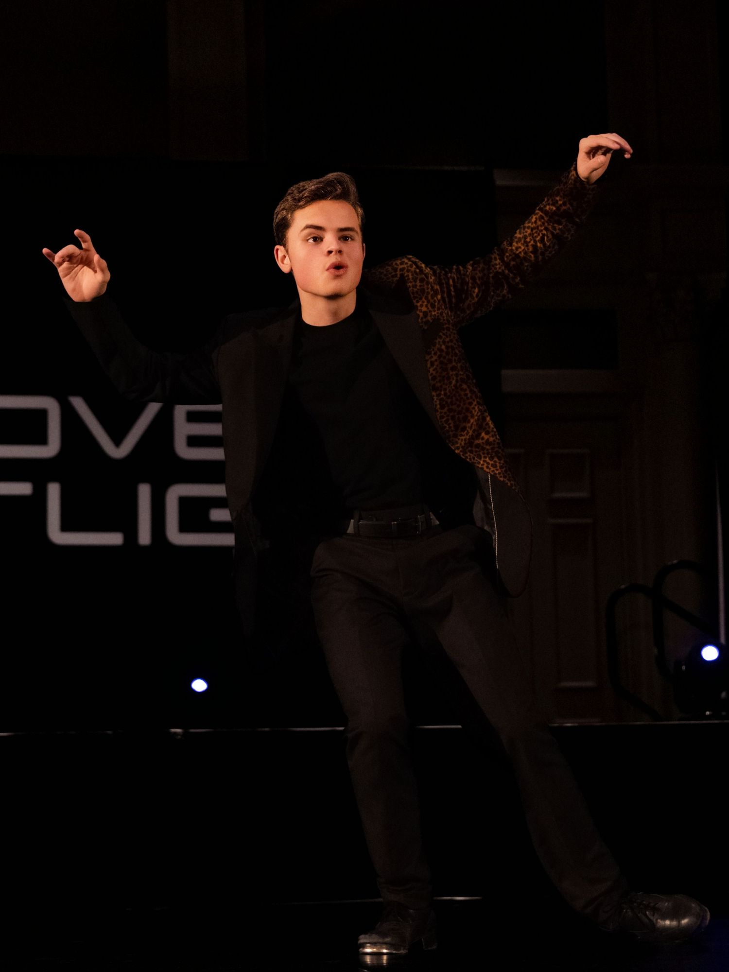 A performer in all black attire strikes a dramatic dance pose on a dark stage with illuminated text visible in background.