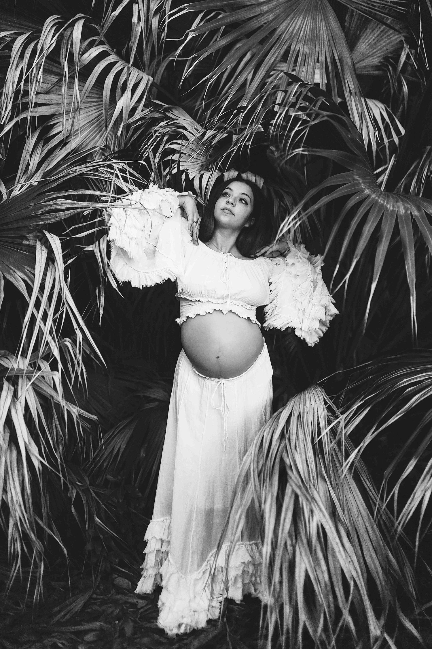 Black and white maternity photo of pregnant woman in white lace dress among tropical palm leaves.