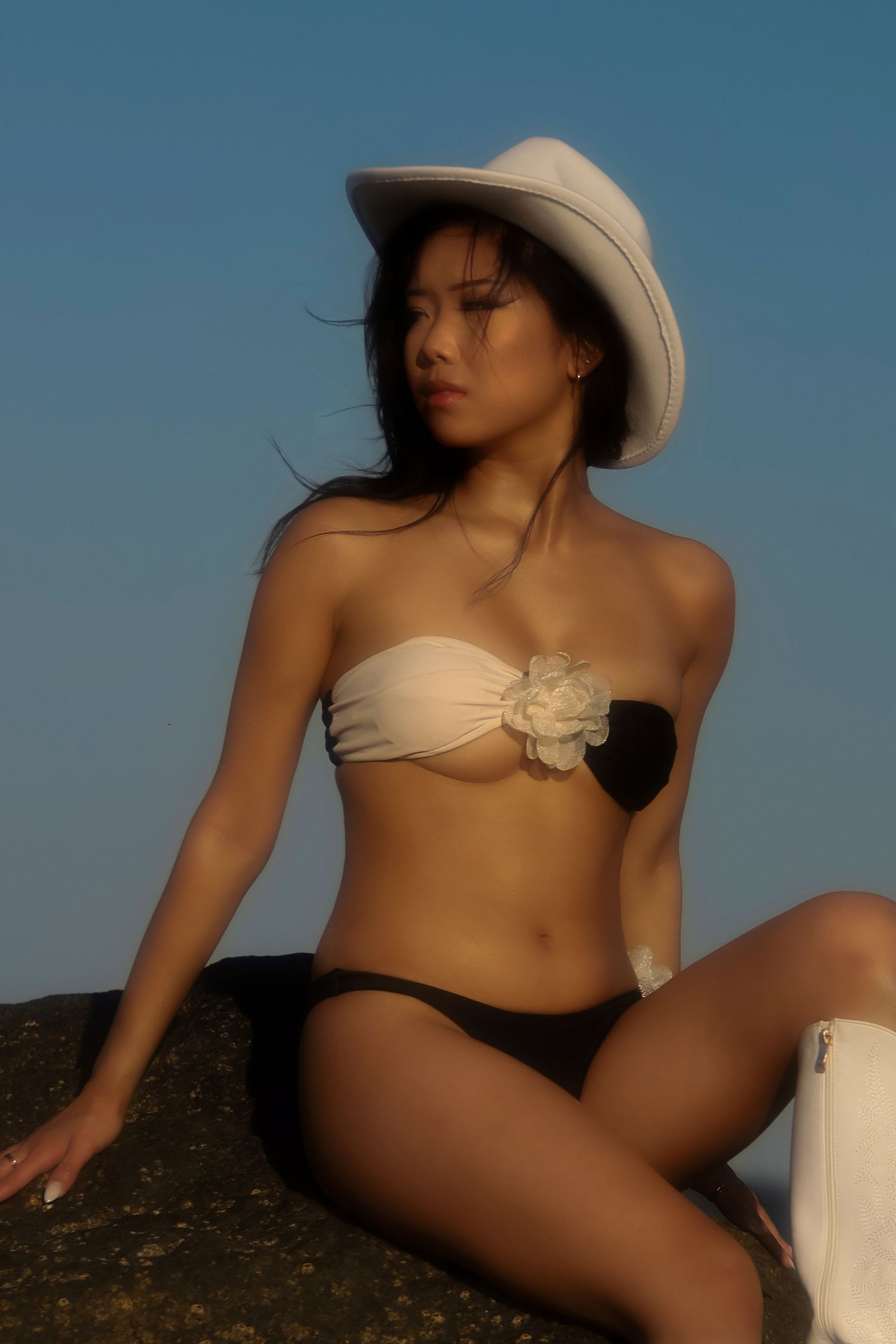 Figure in beige strapless bikini top and black bottom poses on rock at sunset wearing white sun hat.
