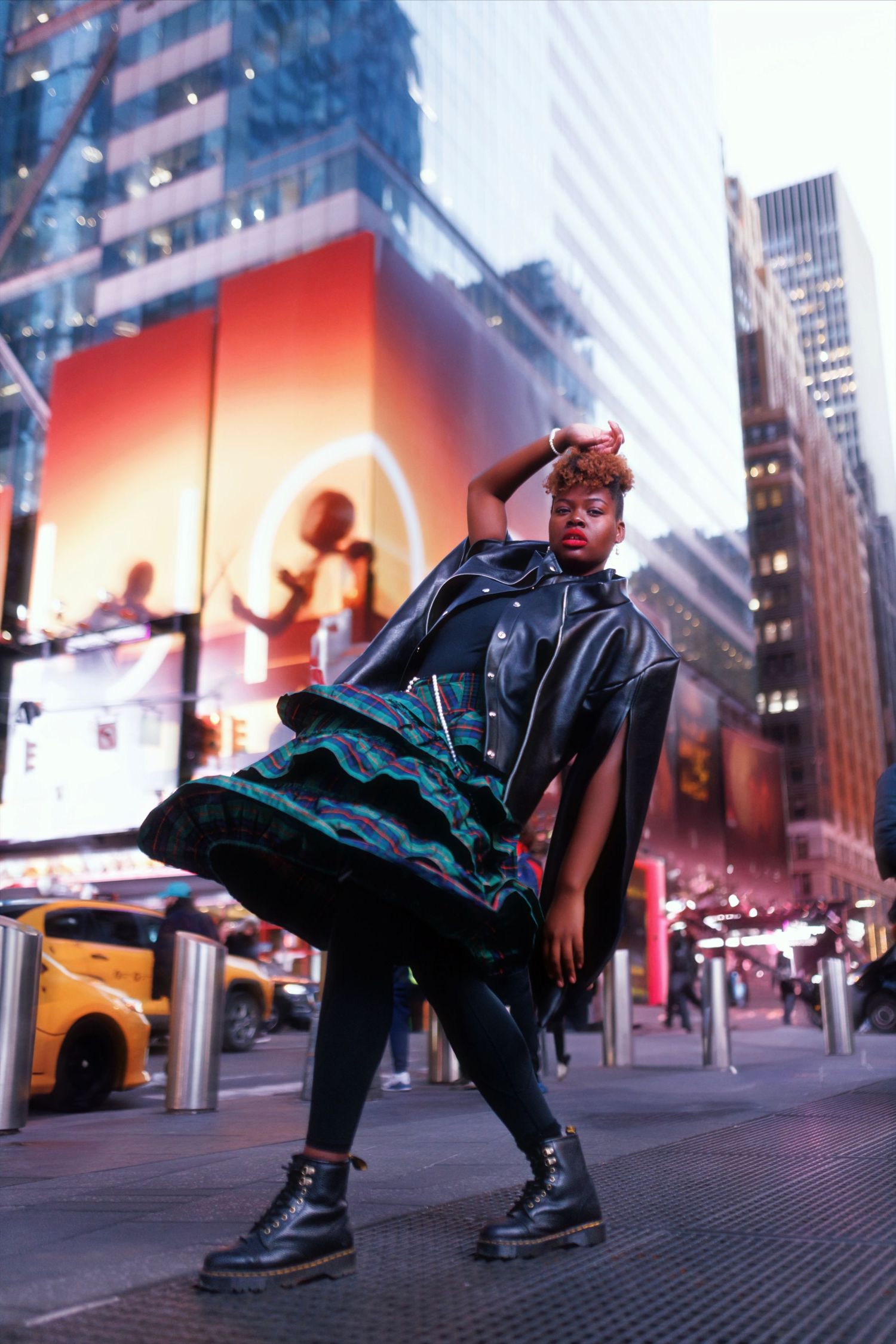 Fashion model poses stylishly in black outfit on Times Square street with illuminated billboards in background.
