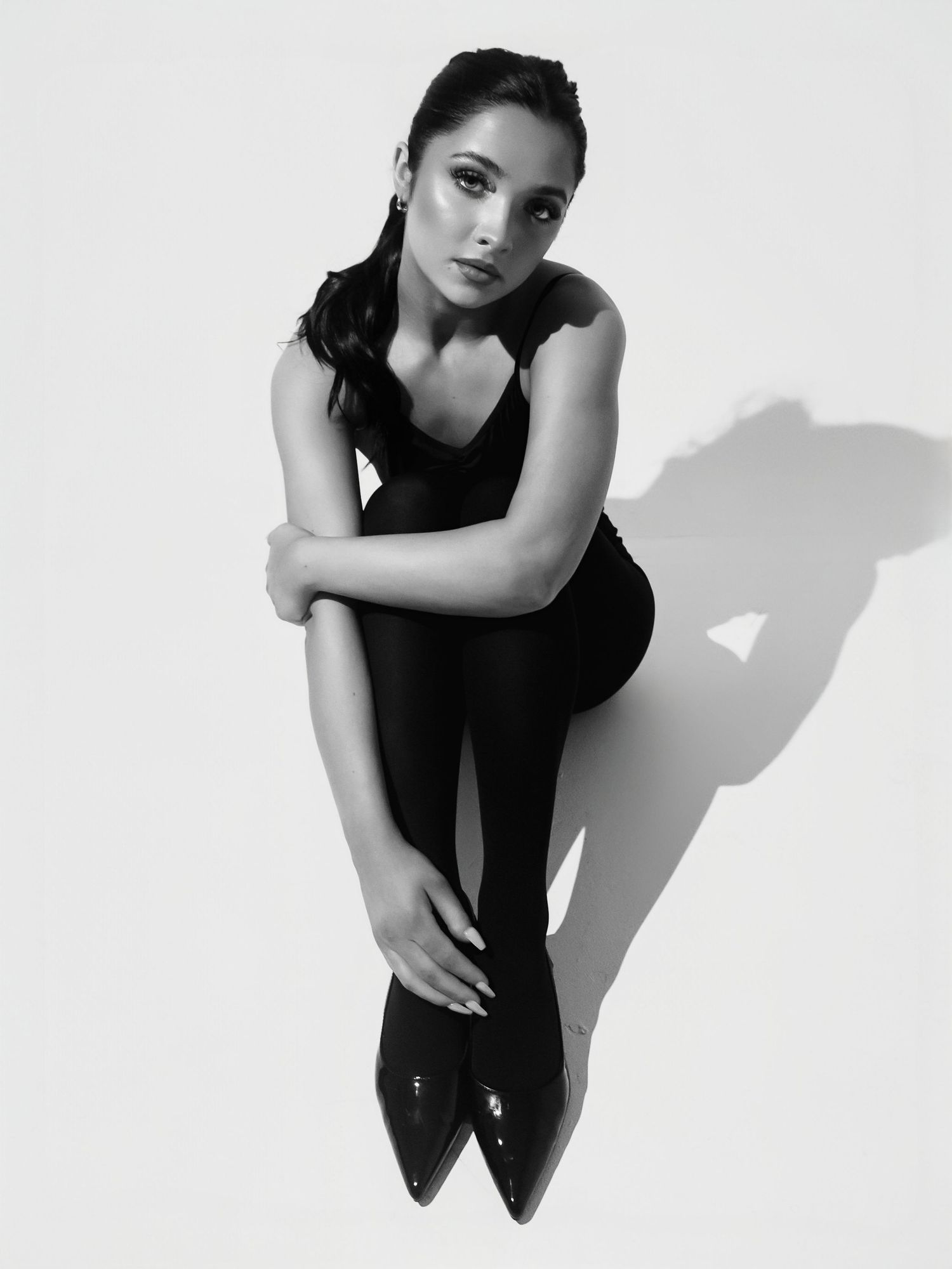 Black and white fashion portrait of a model in dark attire striking an artistic pose against a plain background.