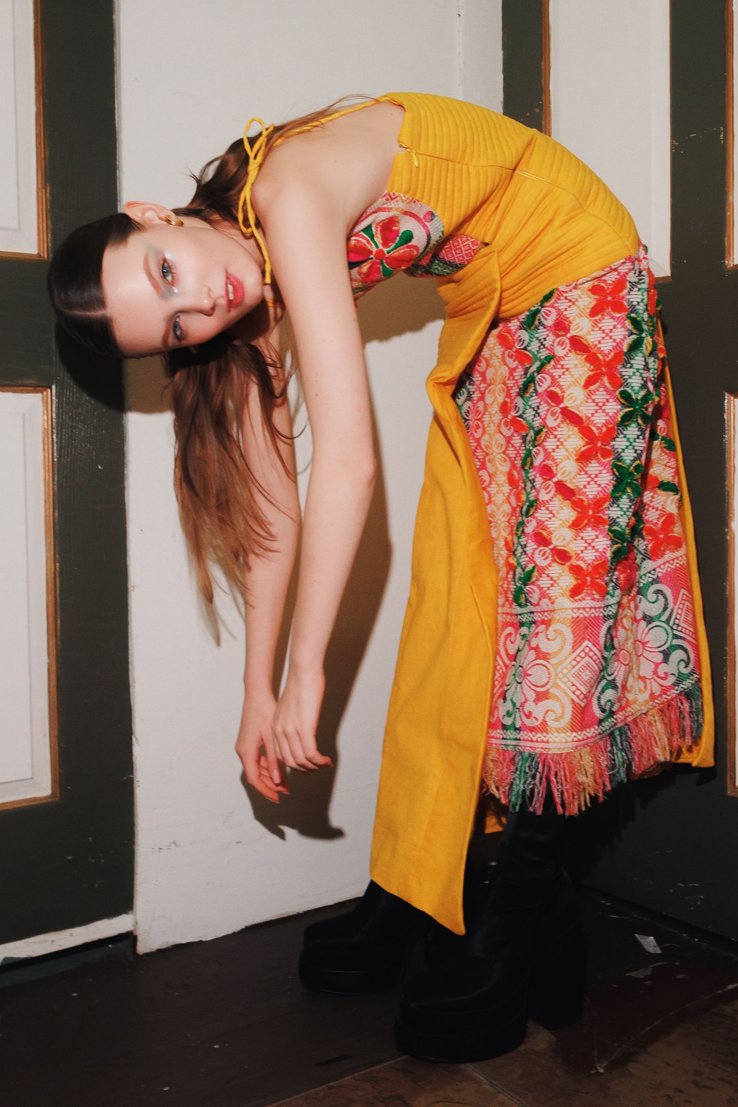 A person in a yellow outfit and colorful patterned wrap bends forward dramatically against a white wall.