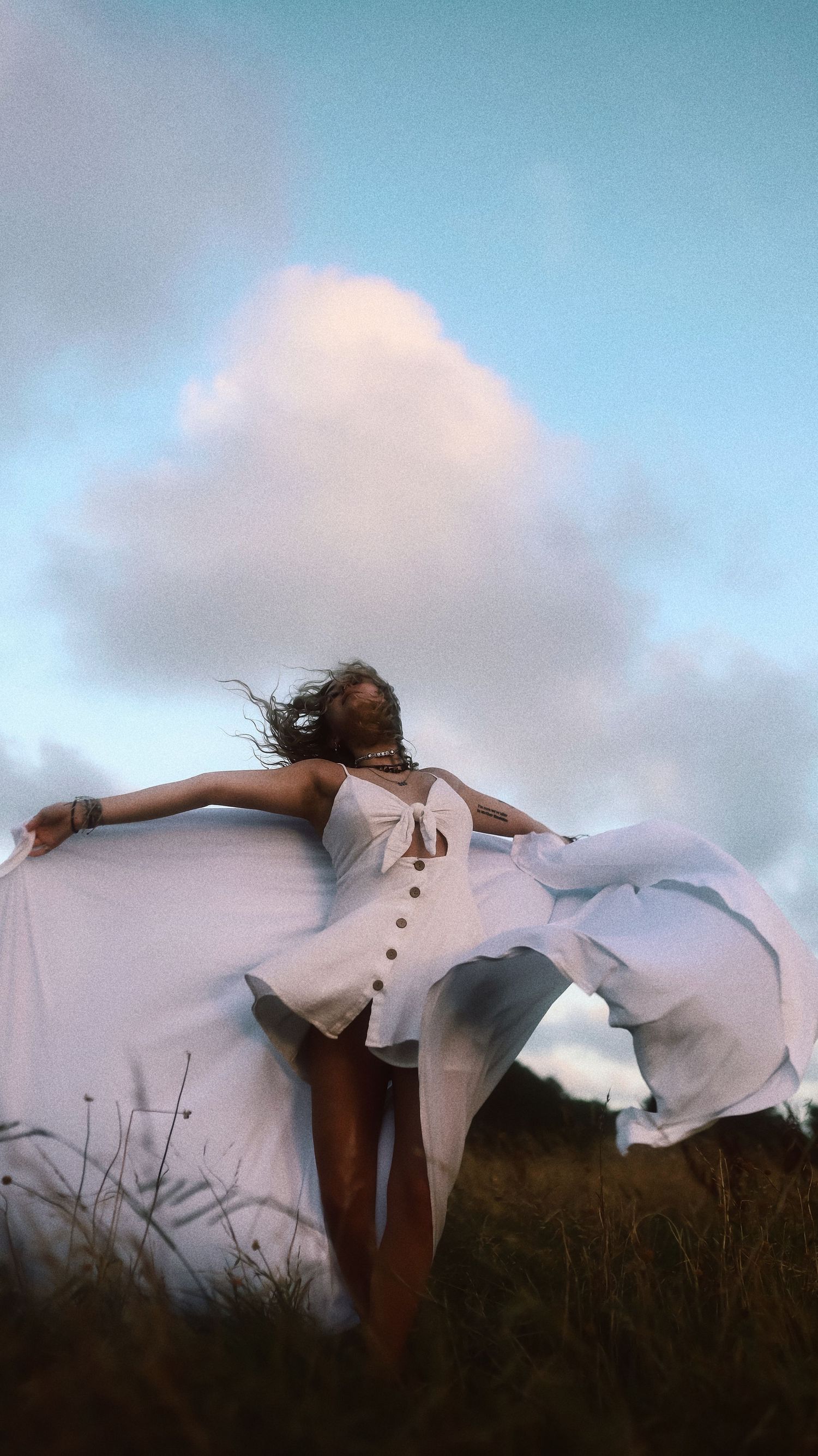 A person in a flowing white dress stands in a field with arms outstretched against a cloudy blue sky.