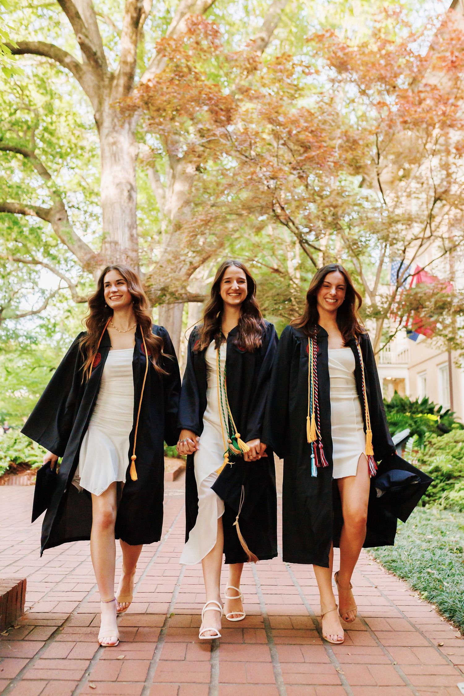 Three graduates in matching black gowns and white dresses walk together on a brick path surrounded by autumn trees.