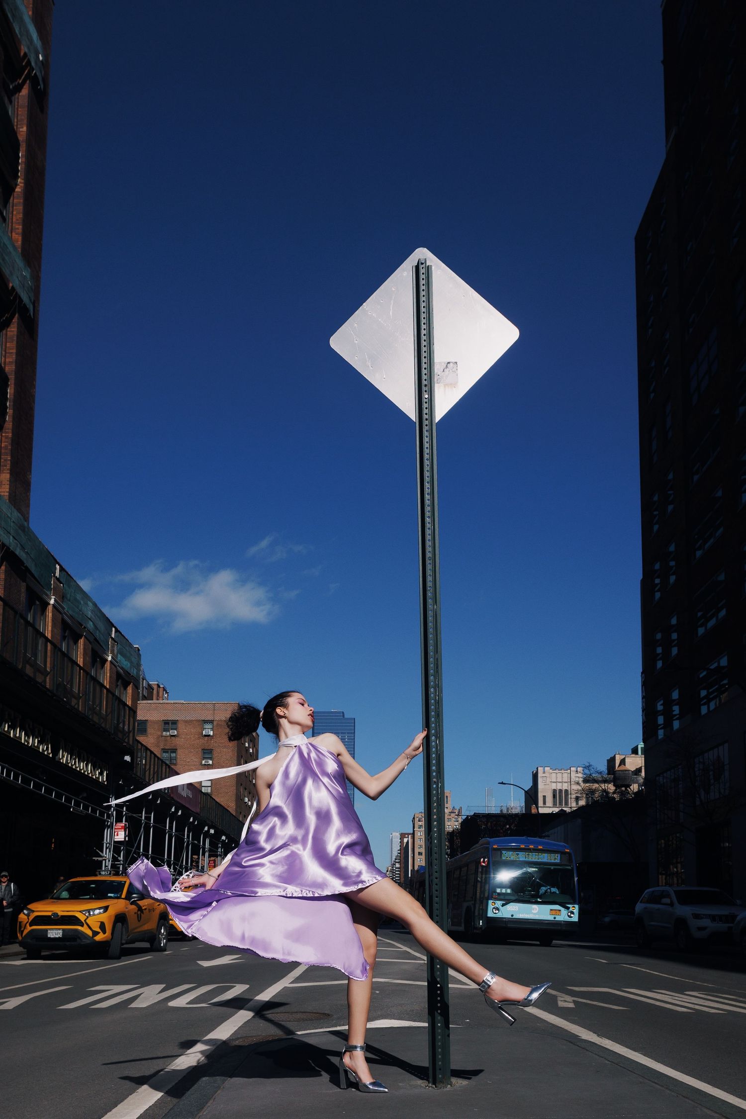 A dancer in flowing purple fabric performs graceful movements on a city street with yellow taxis visible in the background.