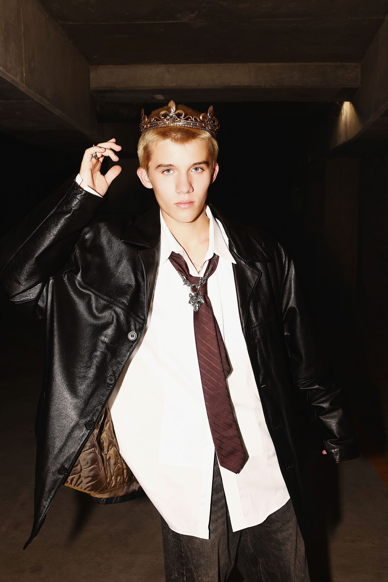 Someone wearing a leather jacket, white shirt, loose tie and gold crown poses in a dimly lit setting.